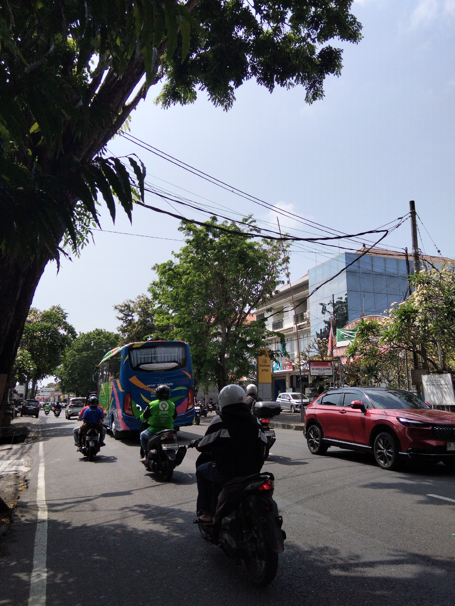 The image depicts a street scene in Denpasar, Indonesia, characterized by a moderately busy road under a clear sky. In the foreground, several motorcycles are visible, with riders wearing helmets. One motorcycle in the immediate foreground has a rider wearing a dark jacket and a white helmet. Behind this, another motorcycle is carrying a rider in a bright green jacket with the "Gojek" logo, suggesting a ride-hailing service. Further back, a large blue bus with colorful graphics is traveling on the road. A red SUV is positioned on the right side of the road, also moving.
The environment includes mature trees lining the street, casting shadows on the asphalt. Buildings are present on both sides of the road, with one building on the right featuring a modern glass facade. Power lines are strung overhead, typical of urban infrastructure. The overall impression is of a sunny day in a developed urban area.