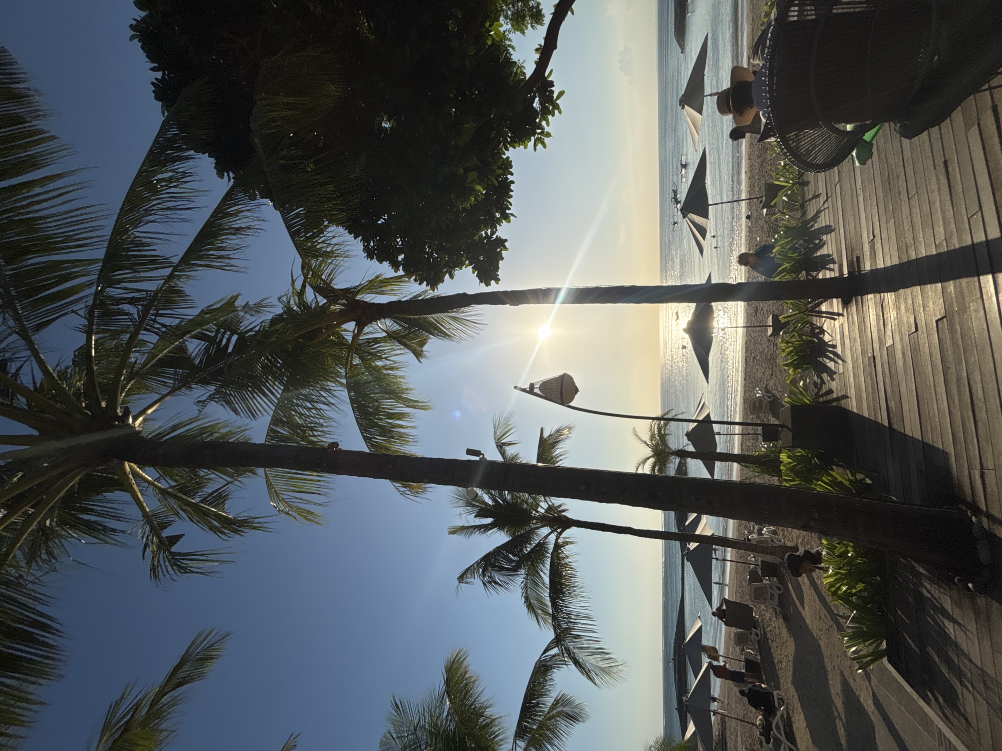 The image depicts a beach scene in Kuta, Indonesia, bathed in the warm glow of the setting sun. Palm trees frame the view, their fronds casting long shadows across a wooden boardwalk in the foreground. Beyond the boardwalk, beach umbrellas dot the sandy shore, providing shade for scattered beachgoers. The ocean stretches out to the horizon, with gentle waves lapping at the shore and a few boats visible in the distance. The sky is a clear, vibrant blue, transitioning to a softer hue near the horizon where the sun is setting. The atmosphere suggests a peaceful and relaxed end to the day at this popular Indonesian tourist destination.