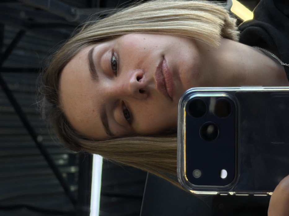 A woman with blond, straight hair, facing forward, holds up a phone taking a selfie. Her features include fair skin, hazel eyes, and medium-full lips. She wears a black top, accented by a silver chain. The phone has a clear case and shows a triple-lens camera system. A reflective surface is visible in the background.