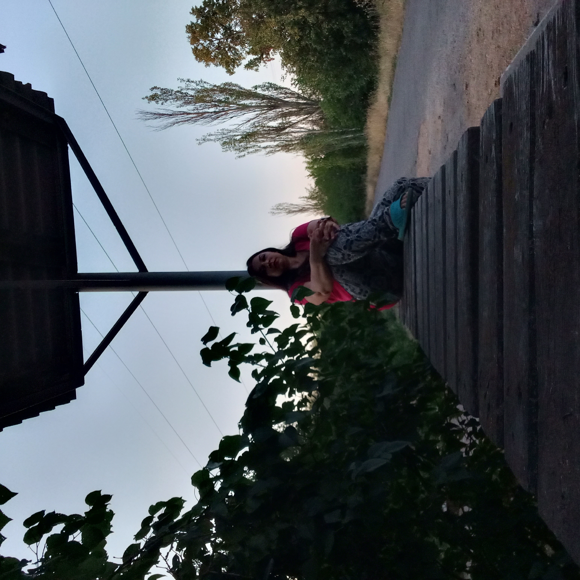 A woman sits on a wooden bench under a shelter with a sloped roof supported by a central pole. The bench is made of thick, dark planks laid horizontally, with the woman positioned on the far right side of the bench, facing forward. She is wearing a bright pink t-shirt and patterned leggings, with her legs crossed and her hands clasped. Her hair is dark and shoulder-length. To the left of the bench, dense green foliage obscures the background. Behind the woman, a paved path runs parallel to the bench, bordered by dry grass and a line of trees. Tall, slender trees with sparse leaves are visible in the distance, along with more dense green trees and bushes. Overhead, power lines are visible against a pale, twilight sky. The scene appears to be outdoors in a park or recreational area, possibly on the outskirts of the city of Berezhynka, Ukraine, given the context. The overall lighting suggests it is either early morning or late evening.