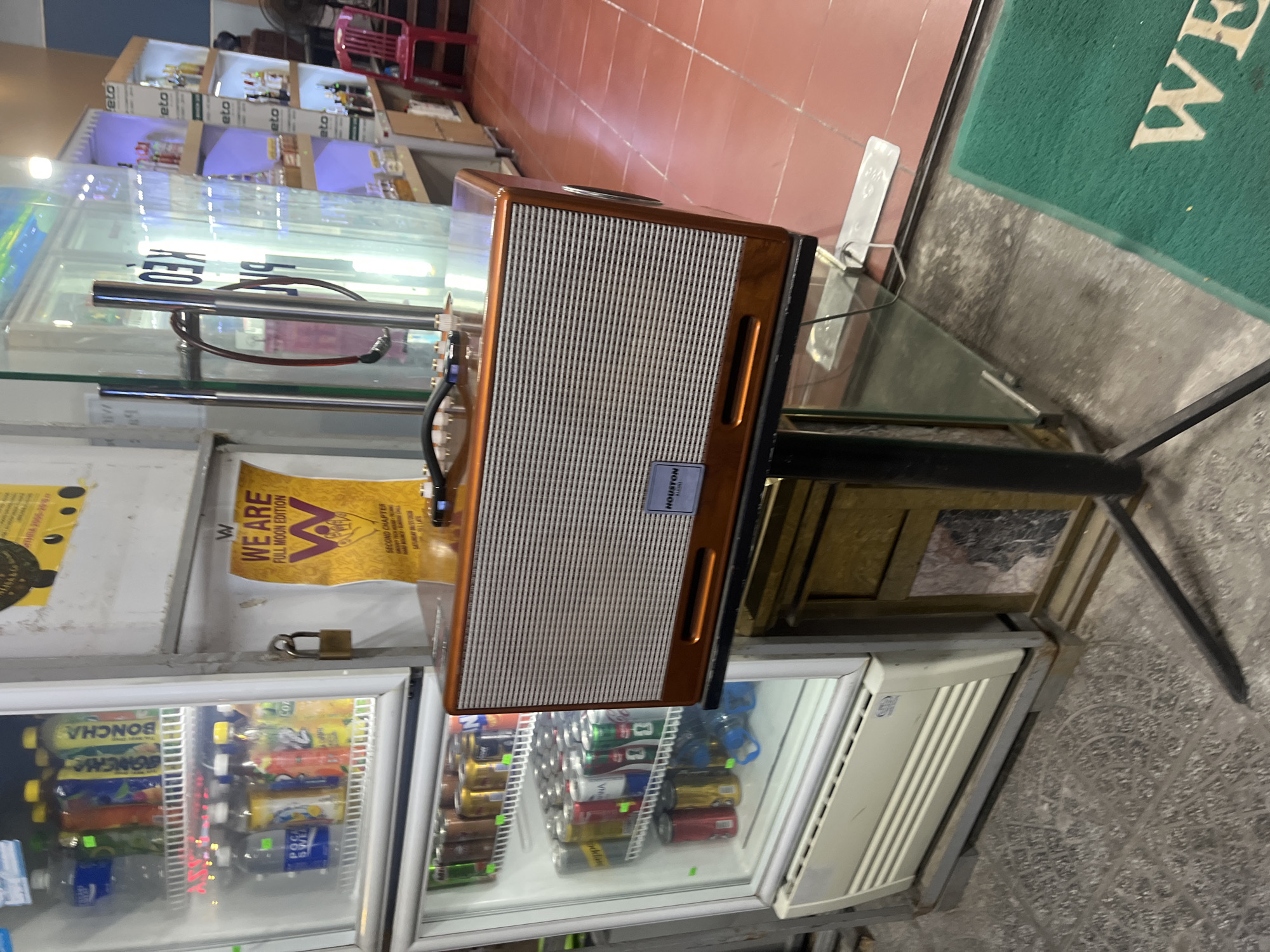 The image captures an outdoor scene, likely a storefront in Đà Nẵng, Vietnam. A white commercial refrigerator, filled with various bottled drinks, is prominently featured on the left. To the right of the refrigerator, a vintage-style orange speaker sits on a stand. Behind the speaker, a poster with the text "WE ARE FULL MOON EDITION" is visible. Further to the right, shelves are stocked with bottles, suggesting a retail environment. The flooring consists of red tiles and gray concrete. The overall impression is that of a shop or convenience store.