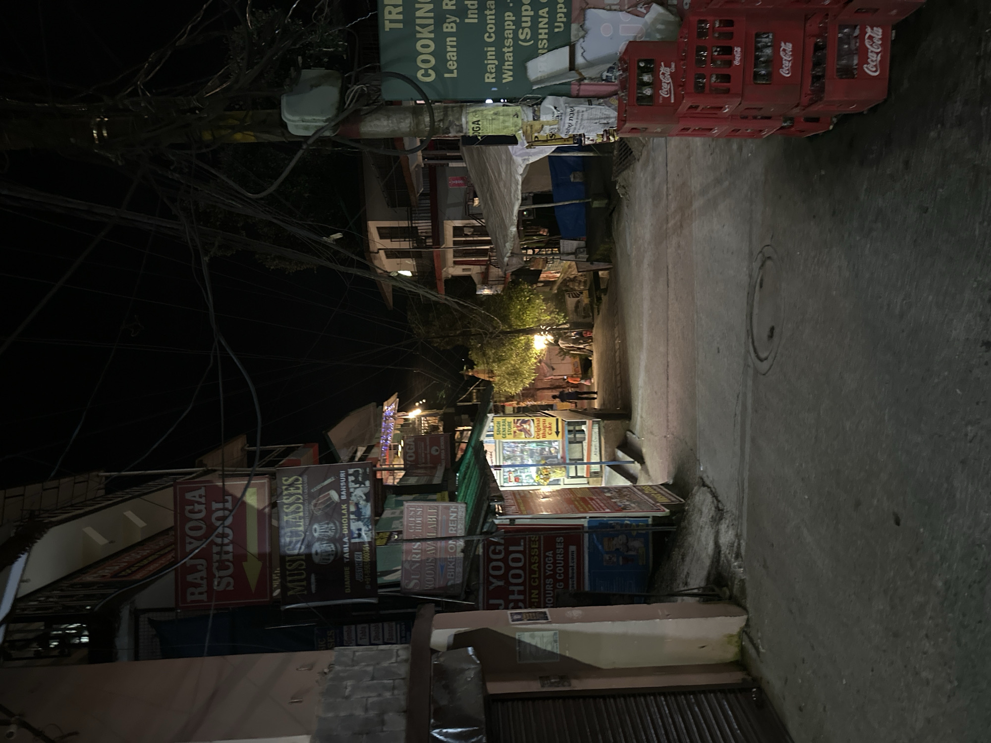 A narrow street in an urban setting is depicted at night. The street is paved and appears to be illuminated by artificial light sources, casting shadows. Buildings line both sides of the street, displaying a variety of signs for businesses such as a yoga school, music classes, and a general store. Several of these signs are illuminated.
On the left side of the street, a building features a prominent red sign for "RAJ YOGA SCHOOL" with a yellow arrow pointing left. Below it, a dark sign advertises "MUSIC CLASSES" with images of musical instruments. Further down, another red sign indicates "J YOGA SCHOOL" with details about classes and courses.
On the right side of the street, a white tent structure is visible, partially covering a pathway. A tall utility pole covered in a dense network of electrical wires stands prominently. Various posters and flyers are affixed to the pole. At the bottom right of the image, several red Coca-Cola crates are stacked, filled with bottles.
Two individuals are visible walking in the distance down the street. The overall atmosphere is that of a commercial area at night, with active businesses and street life.