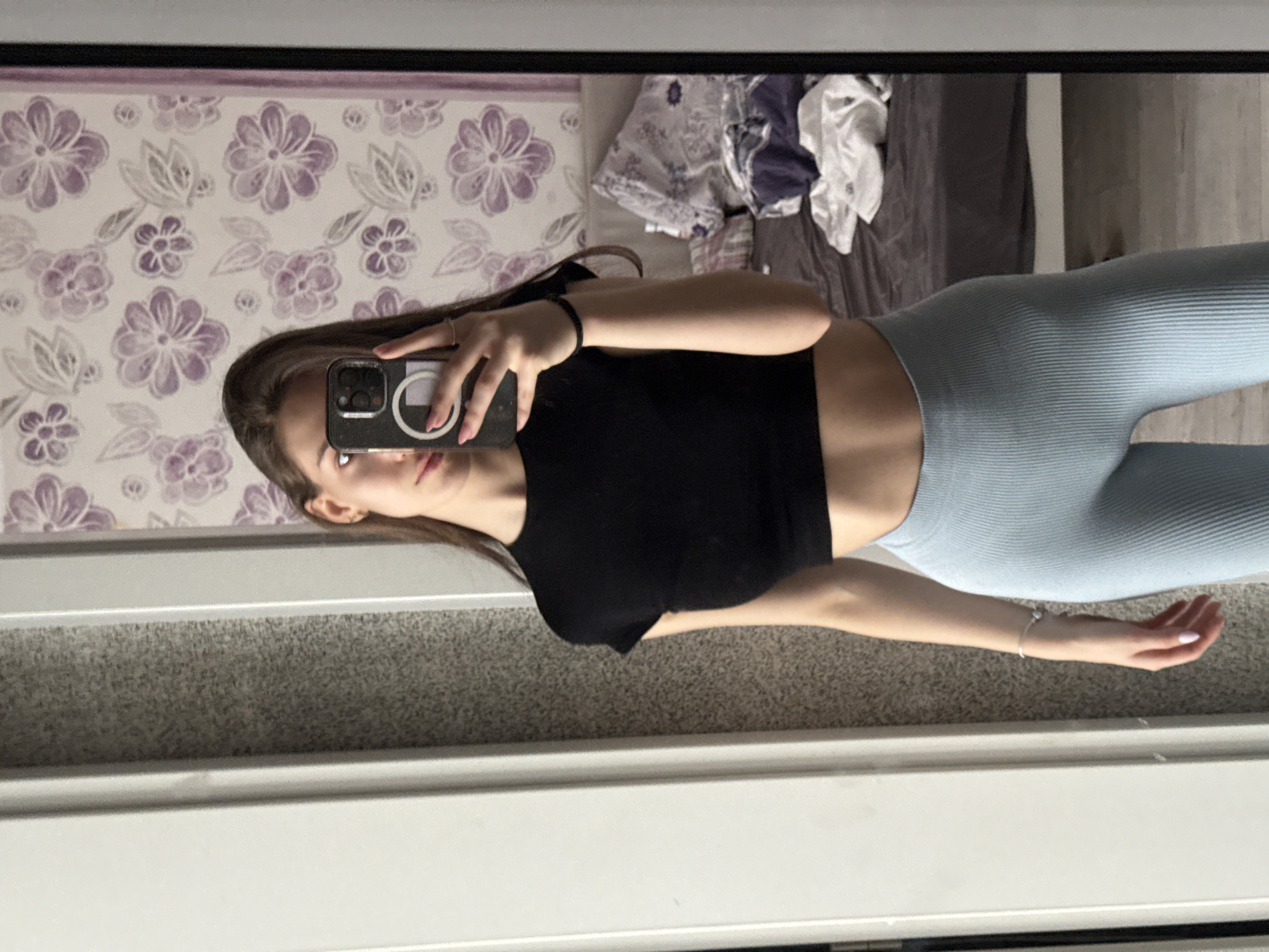 A young woman is taking a selfie in front of a mirror. She is wearing a black crop top and light blue leggings. Her hair is long and brown. She is holding a phone in her right hand. The background includes a wall with a floral pattern, a bed with clothes on it, and what appears to be a section of a door frame or trim. There is no information to indicate that the location is Sofia, Bulgaria.