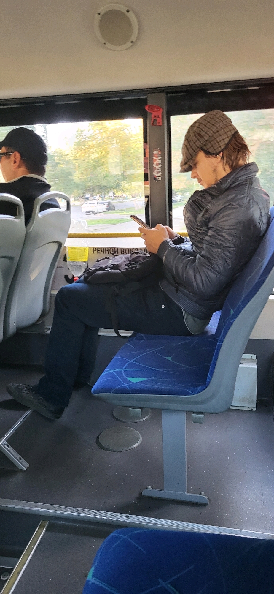 A young man wearing a plaid flat cap and a dark, puffy jacket is seated on a blue patterned bus seat, looking down at his smartphone, which he holds in both hands. He is wearing dark pants and black sneakers. The bus appears to be in motion, with trees and a street visible through the window. A sign with Russian text, reading "РЕЧНОЙ ВОКЗАЛ" (River Terminal), is partially visible behind him. Another passenger is visible in the foreground, wearing a black cap and dark clothing. The interior of the bus is functional, with grey seats and a dark floor. The lighting suggests it is daytime. The specific location in Oskemen, Kazakhstan is not identifiable from the image.
