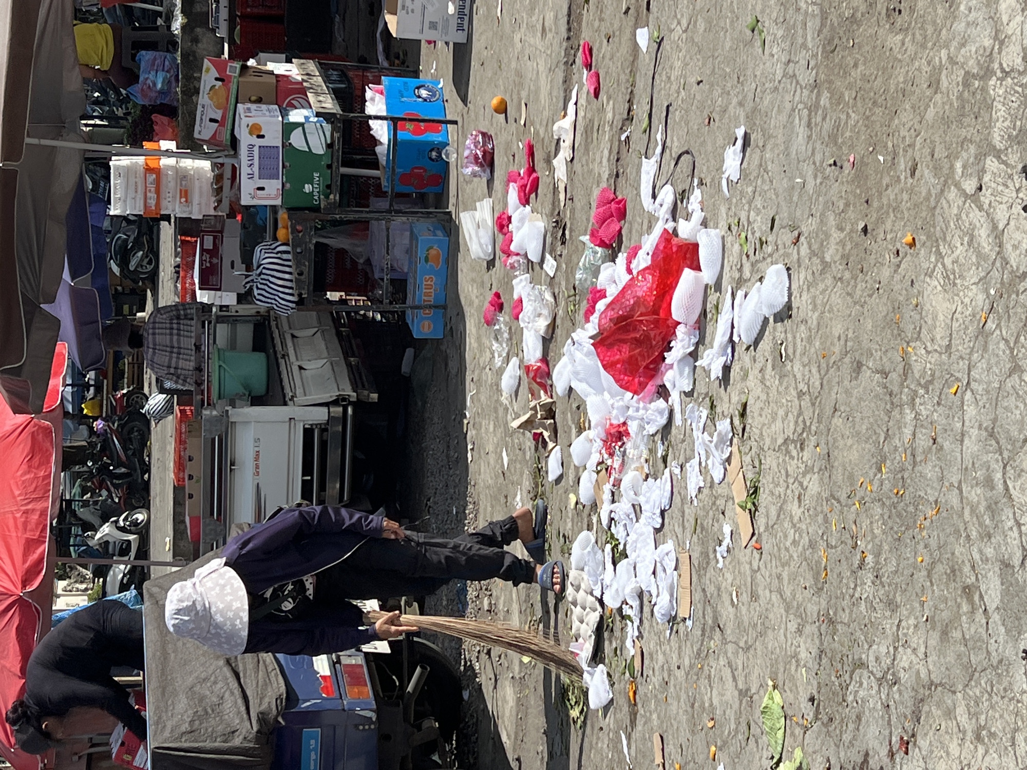 A person wearing a white bucket hat, dark jacket, and black pants is sweeping a concrete surface scattered with various debris, including white and red plastic packaging, food scraps, and small pieces of cardboard. They are using a broom made of straw-like material. In the background, there is a stack of crates and boxes, some labeled with fruit names, stacked near a white vehicle with a visible "Gran Max 1.5" label. Adjacent to the vehicle, there are motorcycles and other market-related items covered by red and brown tarps. The image appears to be taken at Singakerta, Indonesia.
