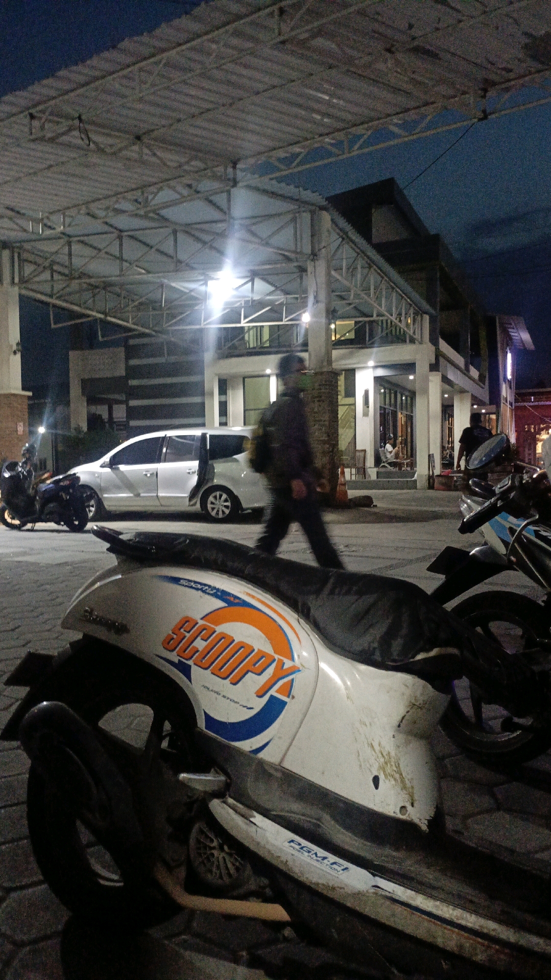At dusk in Ngaru Aru, Indonesia, a white minivan with open rear doors is parked under a metal-roofed structure with exposed beams and lighting. A person walks in front of the minivan. In the foreground, a white scooter with orange and blue "SCOOPY" branding is parked, its rear wheel visible. Other scooters and a person in dark clothing are also present in the background, near a multi-story building with glass windows. The ground appears to be paved with interlocking bricks.