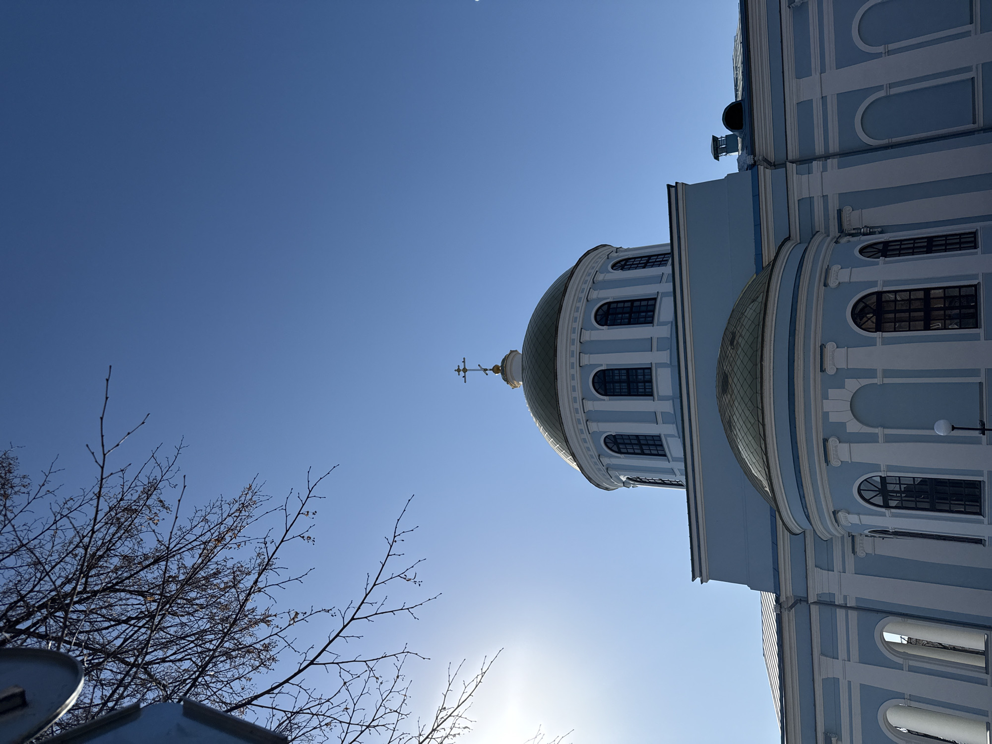 The image shows the exterior of St. Michael's Cathedral in Votkinsk, Russia. The photograph is taken from a low angle, looking upwards at the cathedral's dome and facade against a clear blue sky. Bare tree branches are visible in the lower left corner of the frame. The cathedral is light blue with white architectural details and features arched windows. A golden dome with a cross on top is prominently displayed. The building's structure includes multiple tiers and curves. The sun is positioned behind the cathedral, creating a bright glow.