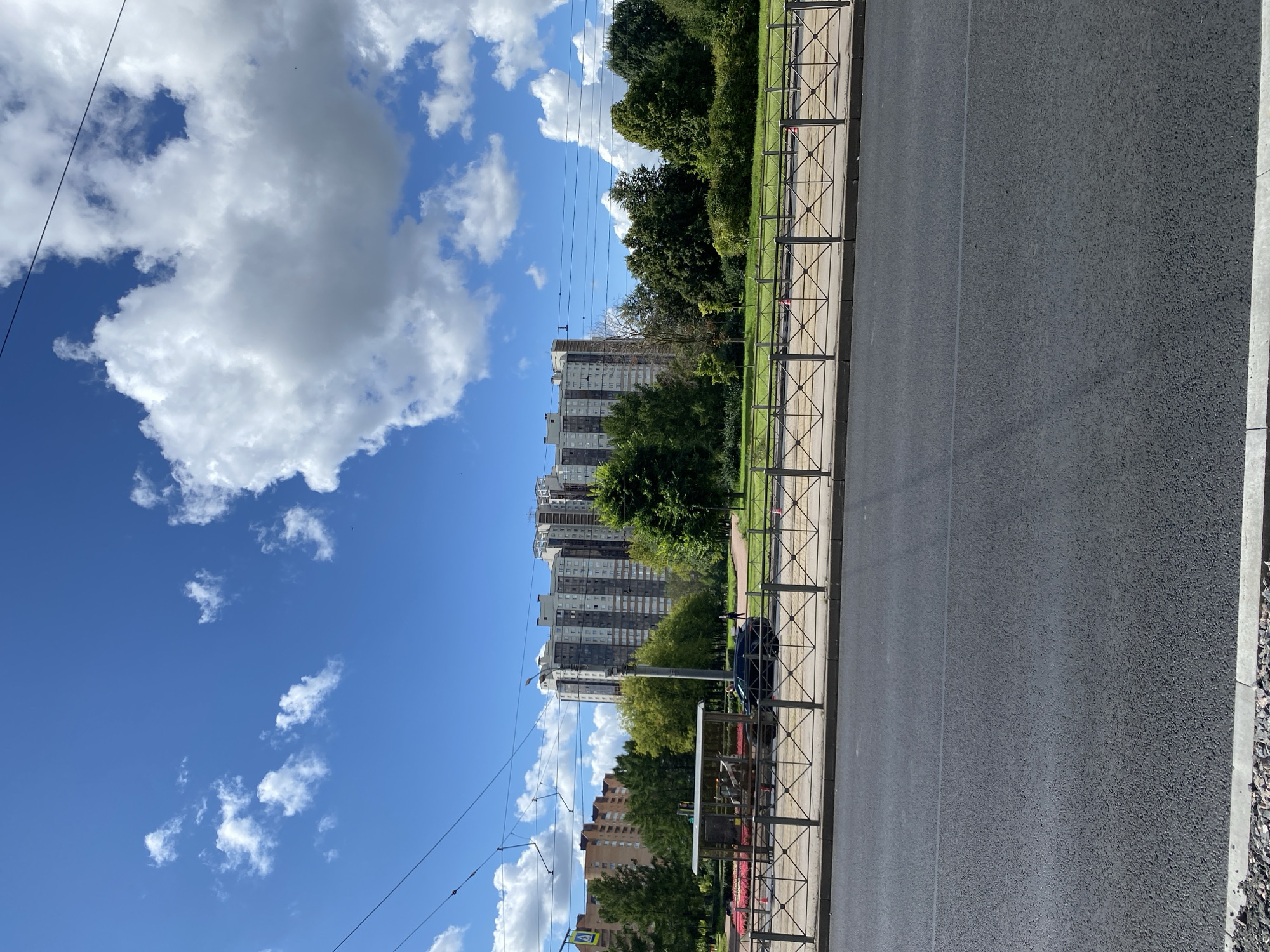 The image depicts a wide asphalt road in the foreground, bordered by a concrete curb and gravel. Behind the road is a pedestrian walkway separated by a black metal fence with a diamond pattern. Beyond the fence, a grassy area with trees and flowerbeds is visible, with a bus stop shelter situated within it. Further in the background, a large, multi-story residential building with a light-colored facade and dark windows dominates the skyline. Several other apartment buildings and lush green trees are also present, suggesting an urban residential area. The sky is a vibrant blue with scattered white cumulus clouds, indicative of clear weather. Power lines are strung across the upper portion of the image. This scene is likely located in Saint Petersburg, Russia, given the architectural style of the buildings and the presence of public transport infrastructure.
