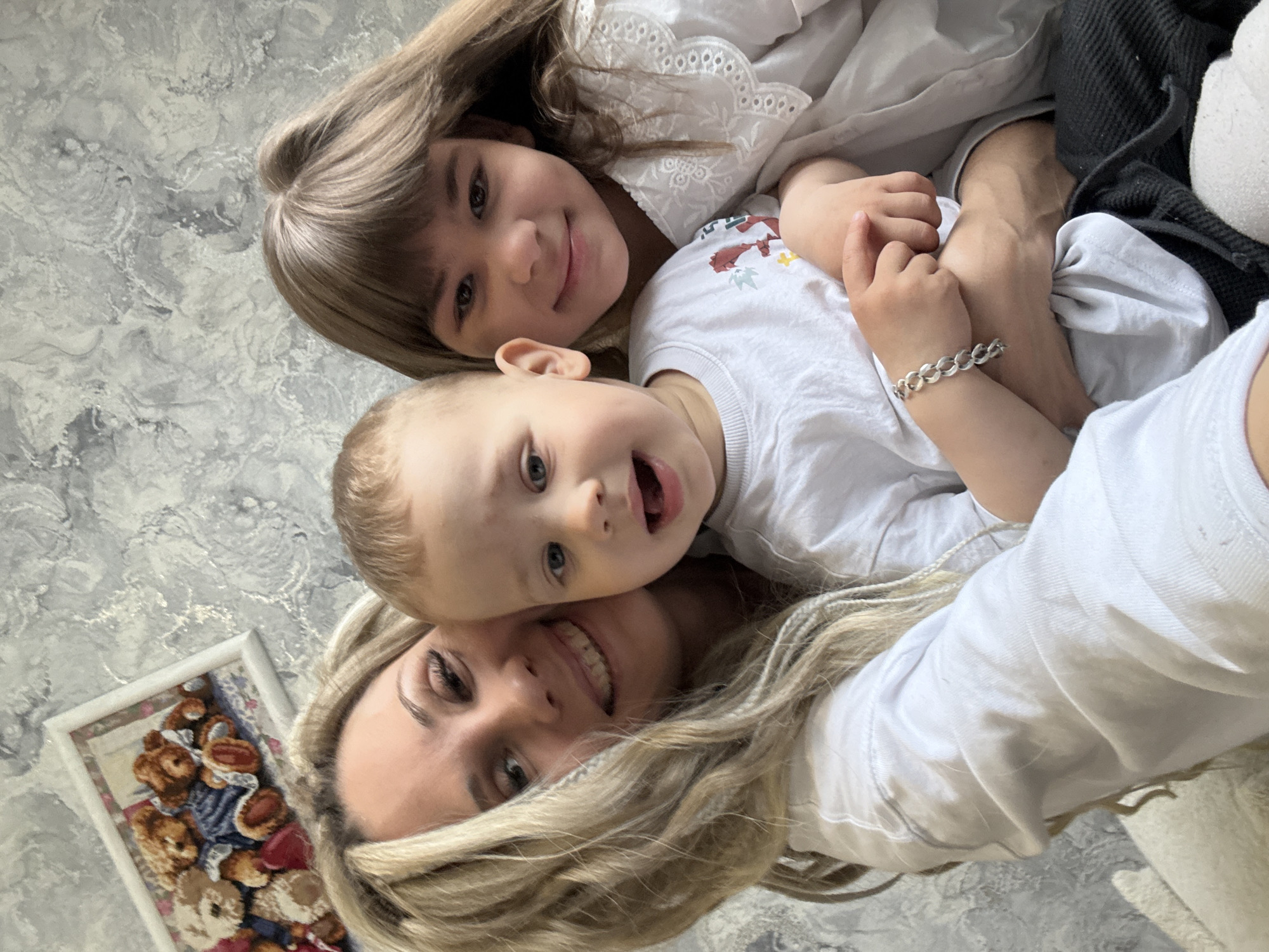 A photograph taken indoors shows a woman with long blonde hair in a white shirt smiling at the camera while holding a young boy with blond hair who is wearing a white t-shirt with a graphic and has his mouth open. Above them is a young girl with long brown hair in a white blouse. The photograph is taken at an angle, with the top of the woman's head towards the bottom left corner and the children oriented towards the top of the frame. The background is a textured wall in gray and white tones. A framed picture of stuffed animals hangs on the wall behind the woman. A portion of black clothing is visible on the right edge of the image. The location is Rusanovo, Russia.
