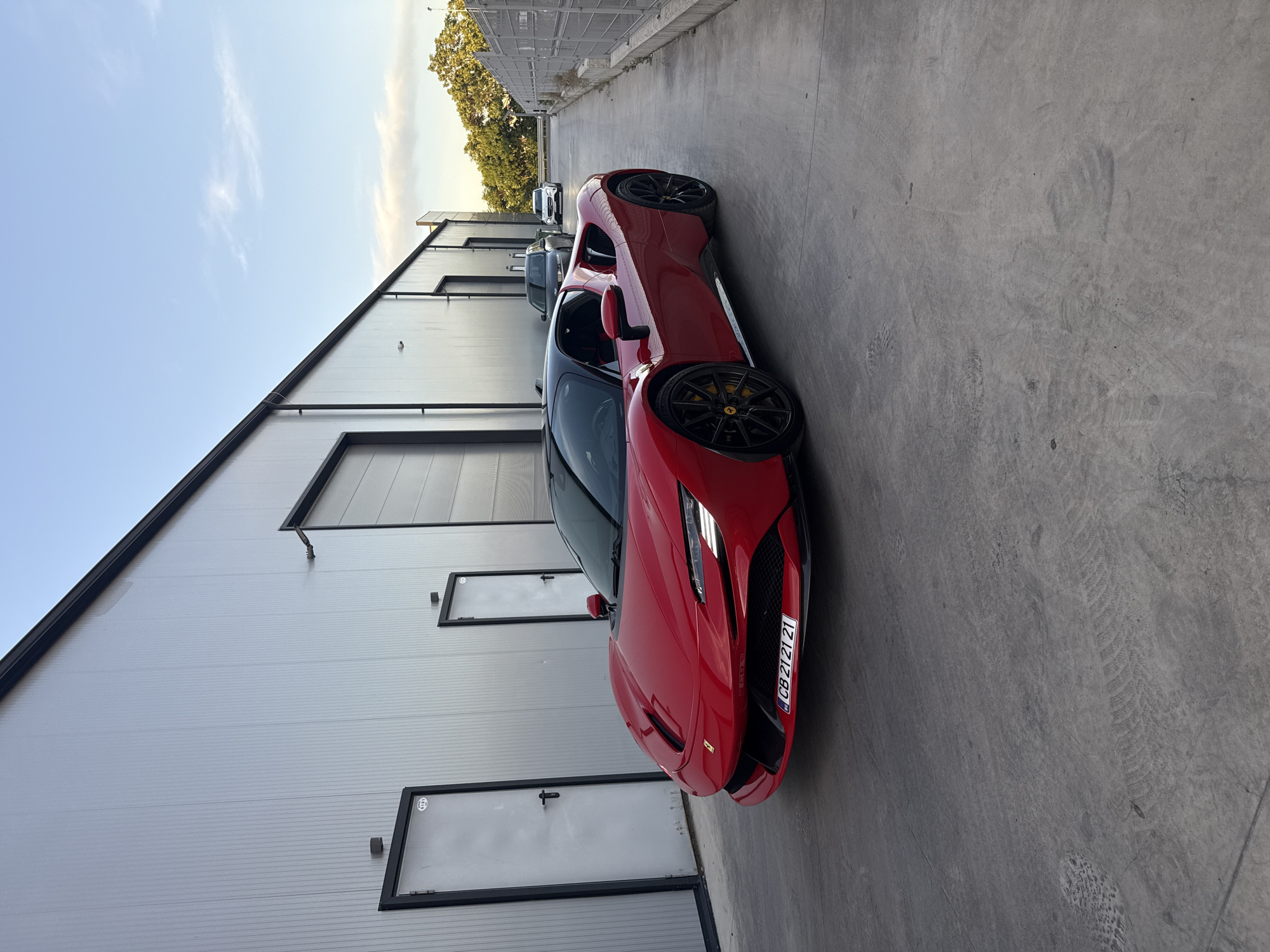 The image depicts a red Ferrari parked in a narrow alleyway in Plovdiv, Bulgaria. The car is positioned diagonally in the frame, facing the viewer. The alley is flanked by a large, grey metal building on the left, with several closed doors. The ground is concrete. In the distance, other parked cars are visible, and the alley leads to an area with trees. The sky is light blue with some light clouds, suggesting it is daytime.