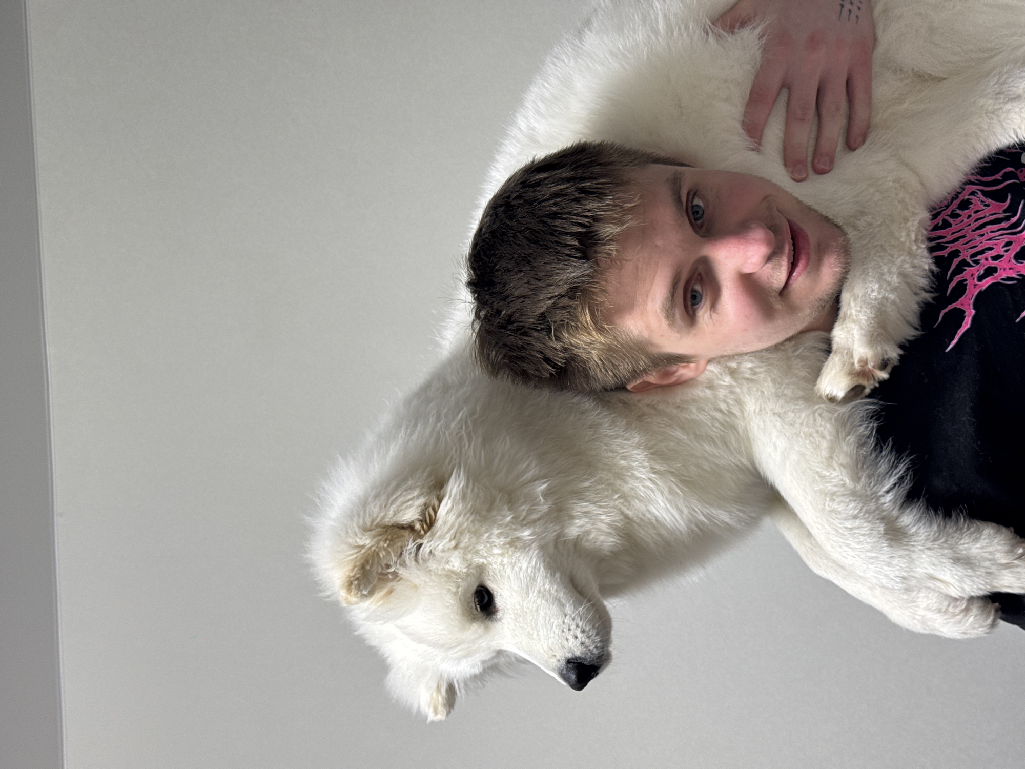 A young man and a large, white, fluffy dog are depicted in a close-up, affectionate portrait. The man, with short light brown hair, blue eyes, and light stubble, is smiling gently. He wears a dark t-shirt with a vibrant pink, intricate graphic design on the right side. His left hand, resting on the dog's back, reveals small, stylized line tattoos on his fingers.
The dog, a breed with thick white fur possibly a Samoyed, has dark eyes and nose, and appears calm and relaxed, nestled close to the man. Its ear, leg, and paw are visible. Both subjects are looking slightly towards the camera, conveying a sense of warmth and companionship.
The background is a plain, light grey surface, suggesting an indoor setting with minimal distractions. The lighting is soft and even, highlighting the textures of the dog's fur and the man's hair. The overall mood is one of comfort and affection between the man and his pet. This scene takes place in Chaikovsky, Russia.