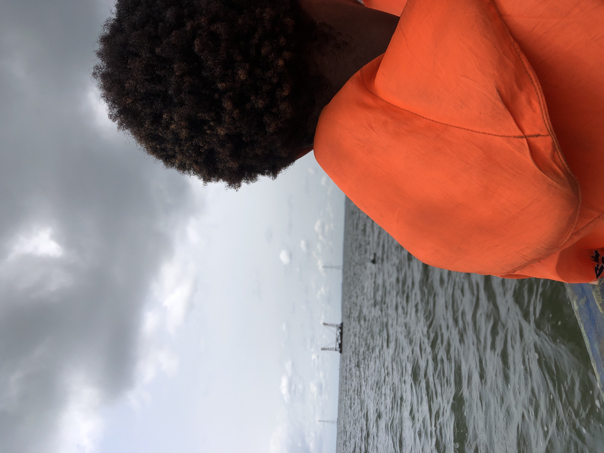 The image, rotated 90 degrees counter-clockwise, depicts a person from behind and slightly to the side, looking out over a large body of water. The person has dark, curly hair and is wearing a bright orange garment, possibly a life jacket, which occupies a significant portion of the right side of the frame. The fabric of the garment shows stitching details and some faint discoloration.
The setting appears to be a boat or a similar vessel, with a dark blue edge visible in the bottom right corner. The main subject of focus for the person is a vast expanse of dark, choppy water, characteristic of a lake, sea, or wide river. In the distance across the water, several thin, vertical structures and one larger, darker structure resembling a platform or building are visible under an overcast sky.
The sky is predominantly grey and cloudy, with some lighter patches indicating breaks in the cloud cover. The overall impression is of a daytime scene with cloudy or stormy weather conditions. No specific activity beyond looking out over the water is discernible. There is no visible text in the image.