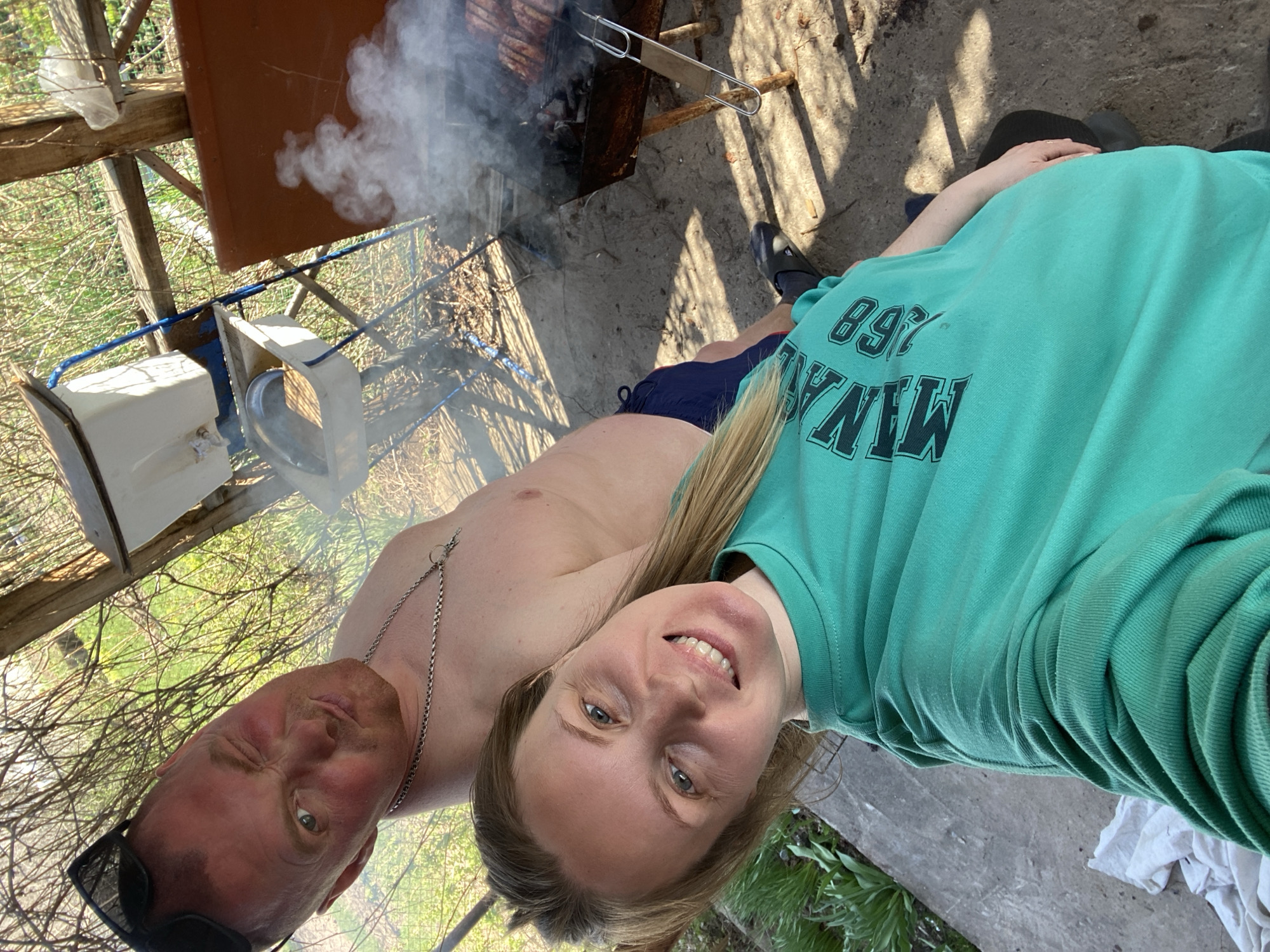 A rotated outdoor selfie shows a shirtless man with sunglasses on his head, a silver necklace, and an apparent sunburn, standing next to a smiling woman in a green sweatshirt. Smoke rises from a barbecue grill in the background. The location appears to be a backyard or outdoor area near a structure with scaffolding and foliage. There are metal objects and containers in the background. The image was likely taken in Chaikovsky, Russia.