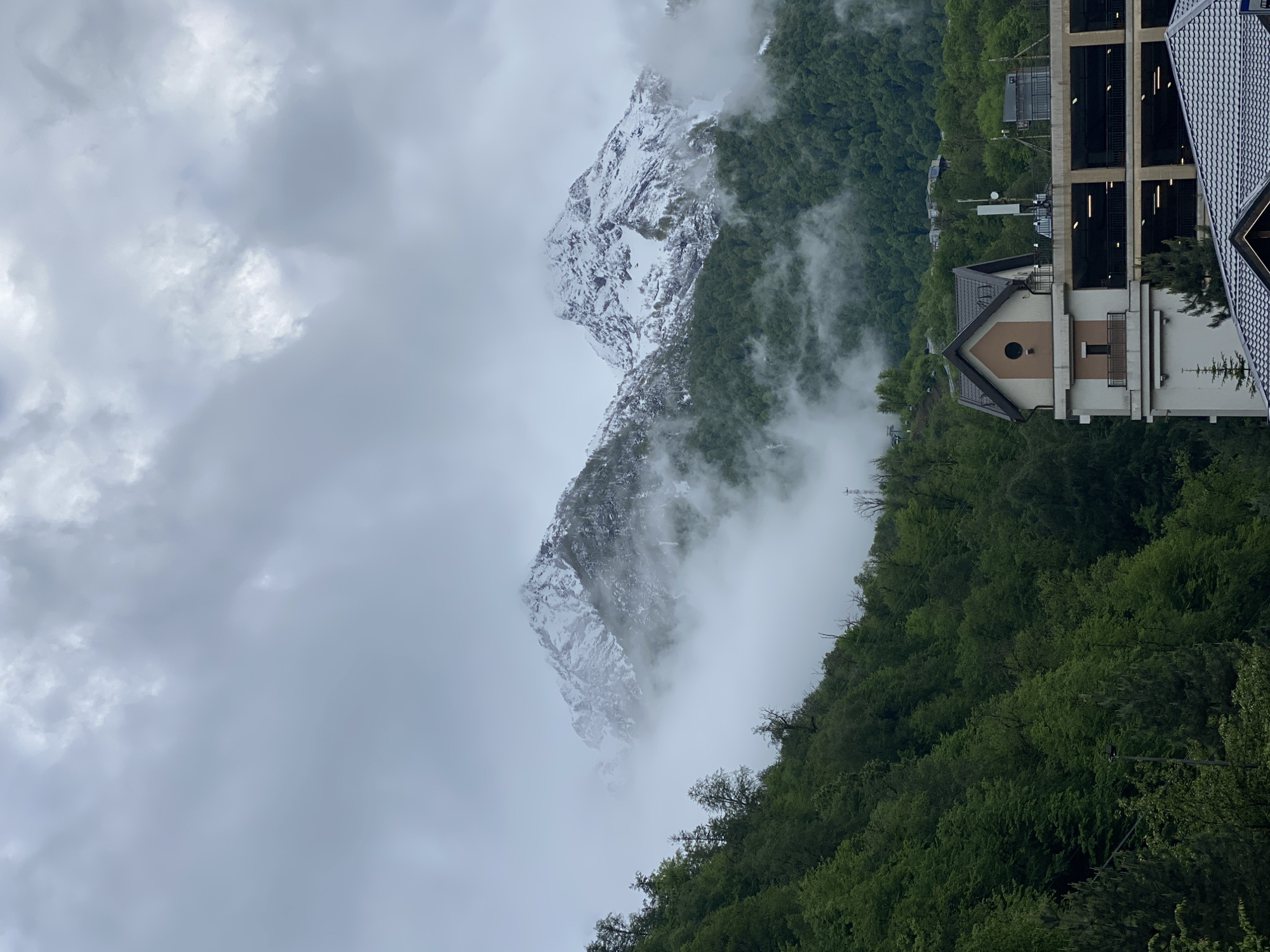 The image depicts a mountainous landscape with a building complex in the foreground, likely located in Estosadok, Russia. The mountains are partially covered in snow and obscured by fog or low-lying clouds. Lush green trees densely populate the lower slopes. The building features a combination of architectural styles, with a lower section having a peaked roof and a round window. Adjacent is a larger, multi-story structure, possibly a hotel or apartment building. The sky is overcast with gray clouds.