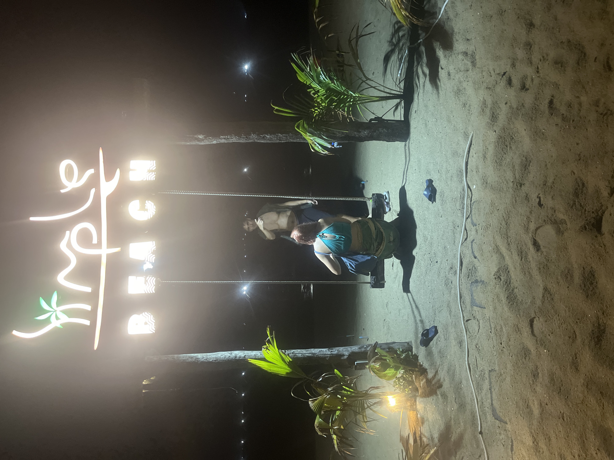 The image depicts a neon sign reading "SIMPLE BEACH" illuminated at night on a sandy beach. A wooden swing hangs from two ropes attached to palm trees. A woman with red hair, wearing a teal top and a patterned skirt, sits on the swing facing away from the camera. A man with dark hair and an open, unbuttoned shirt stands behind her, also facing away from the camera. Tropical plants are visible on the left and right sides of the scene, with a light illuminating the plants on the left. The background shows a dark horizon with faint lights, suggesting a populated area or distant lights of the city of Da Nang, Vietnam. Footprints are visible in the sand in the foreground, and a pair of dark flip-flops rests on the sand near the woman on the swing. A white cord runs across the sand in the foreground.