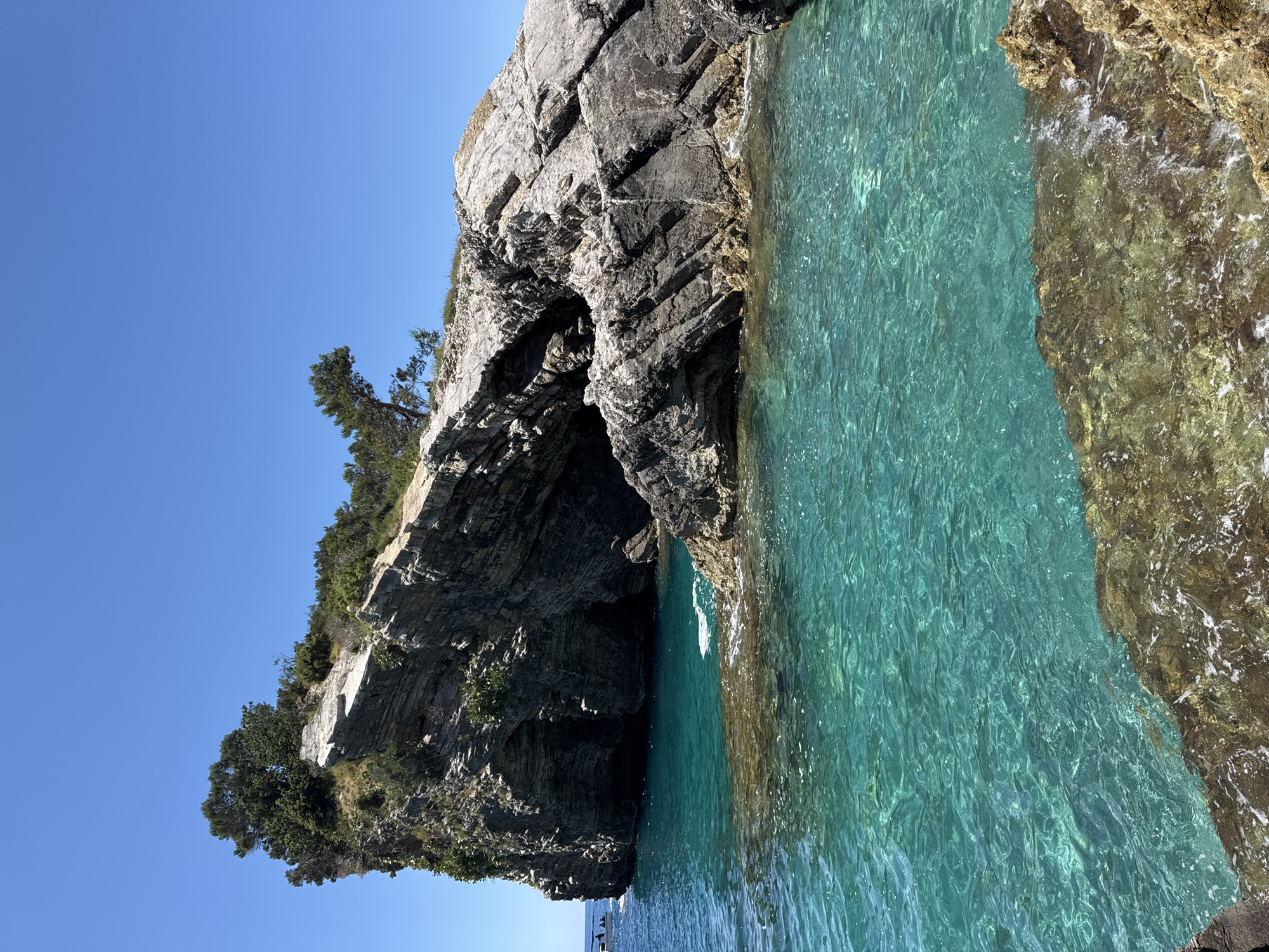 This media file displays a stunning coastal landscape, captured in Budva, Montenegro, and presented with a 90-degree clockwise rotation. The primary subjects are the clear turquoise sea and imposing rocky cliffs under a brilliant blue sky.
The vast expanse of the sky, a deep, unblemished blue, occupies the left side of the frame. To its right, rugged grey and brown rock formations rise vertically, showing distinct layers and jagged textures. These cliffs are adorned with scattered green vegetation, including small trees and shrubs that cling to their surfaces and crown the top. A notable feature is a dark, cavernous opening or archway within the cliffs, visible in the central-right area, suggesting natural erosion by the sea.
The water below the cliffs is exceptionally transparent, exhibiting a striking turquoise to cyan color. Its surface is dappled with ripples, clearly showing the rocky bottom beneath. The transition from water to land reveals darker, wet rocks along the waterline. In the bottom part of the image (which would be the lower left corner if rotated correctly), a small white boat with a few people aboard is visible, hinting at leisure activities like boating or sightseeing along the scenic coastline.
The scene depicts a bright, sunny day with calm weather. The clear sky and crystalline waters evoke feelings of tranquility and natural beauty. There is no visible text in the image.