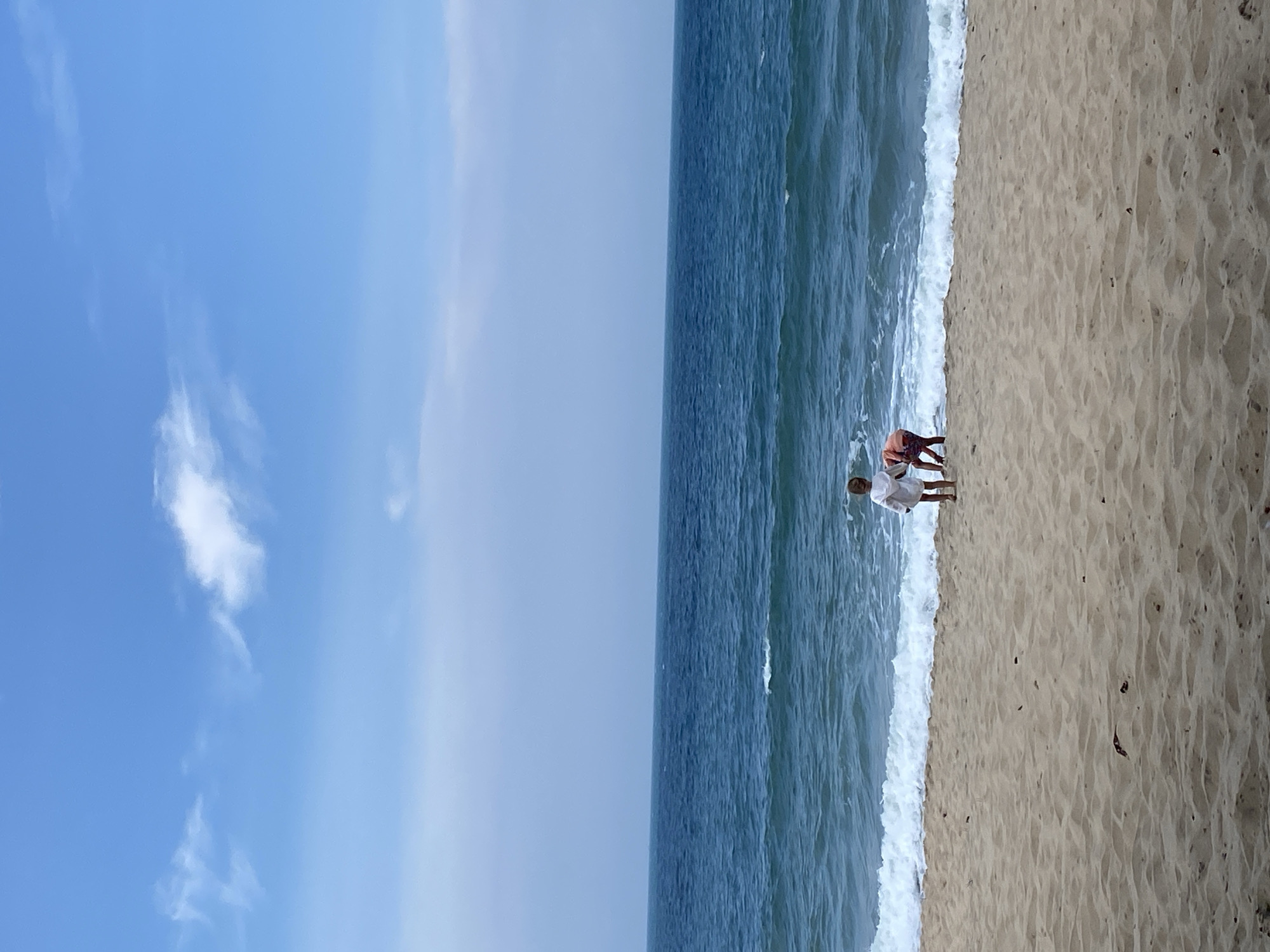 Two young children are on a sandy beach in Phu Thuy Ward, Vietnam, facing the ocean. The child on the left, wearing a white outfit, stands near the water's edge. To the right, a second child in orange shorts and a patterned shirt is partially obscured, leaning down near the waves. The sea is a deep blue with white-capped waves breaking on the shore. The sky above is a pale blue with a few wispy white clouds. The sand is a light tan, showing ripples from the wind or receding water. The overall scene depicts a peaceful day at the beach.
