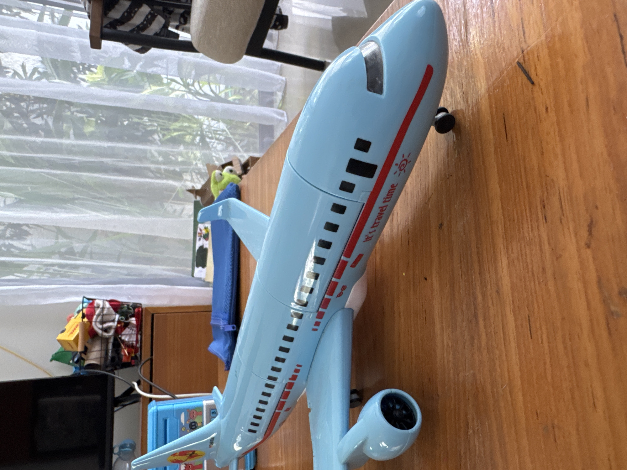 A blue toy airplane sits on a wooden surface. The airplane has black squares and red lines along its body, with the words "It's travel time" printed on it. In the background, there is a window with white curtains, a black metal basket holding various items, a blue object, and a wooden cabinet. The location is not determinable from the image.