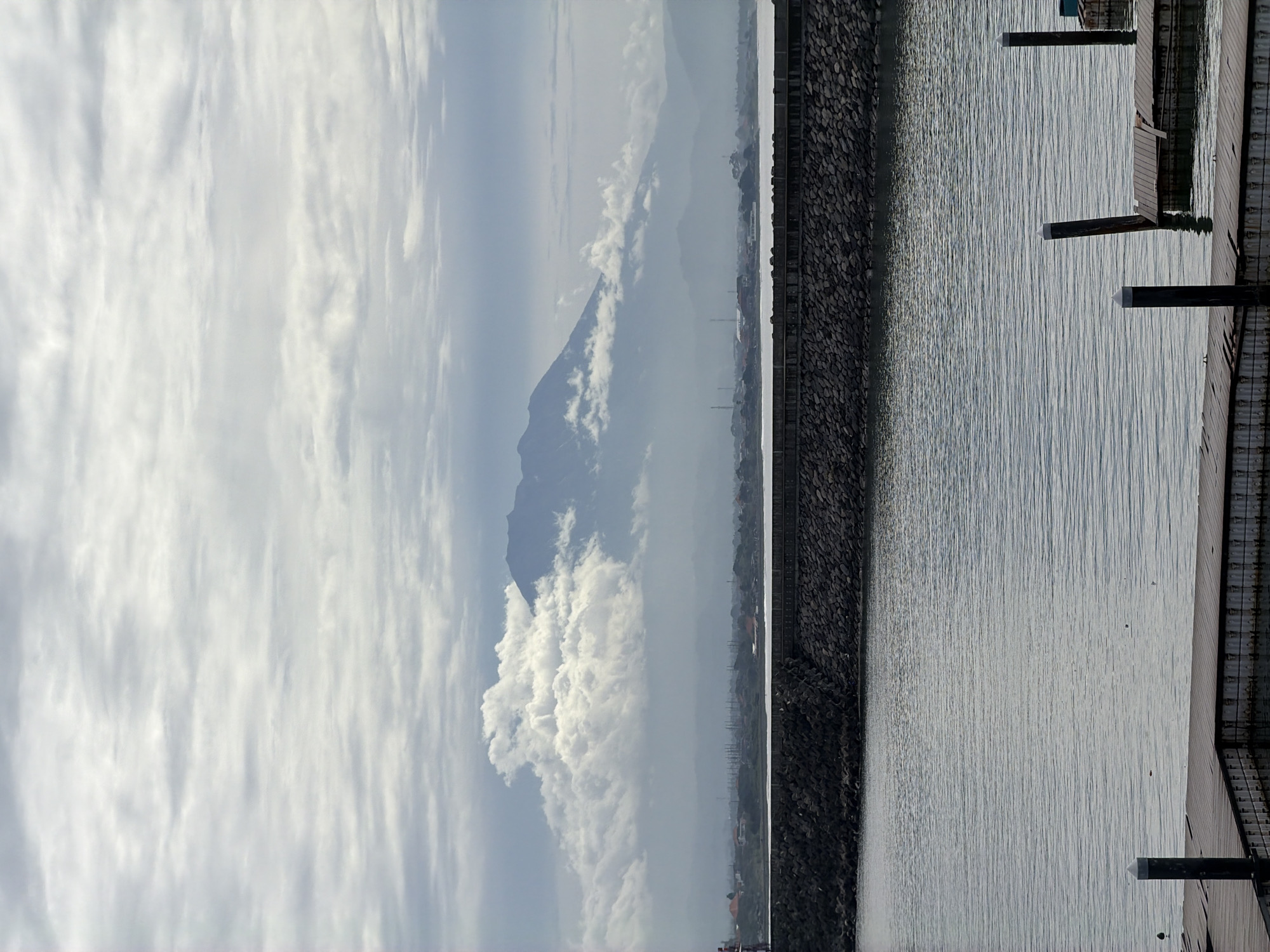 The image depicts a view from a waterfront area, likely in or near Denpasar, Indonesia, suggested by the presence of what appears to be Mount Agung in the distance, obscured by clouds. The foreground consists of a body of water, possibly a bay or harbor, with a stone seawall or breakwater running along the shoreline. A wooden pier or dock extends into the water from the right side of the frame, supported by pilings. The sky is overcast with a mix of light and dark clouds. The scene is static, and no people are visible. The focus is on the natural landscape and the built environment along the waterfront.