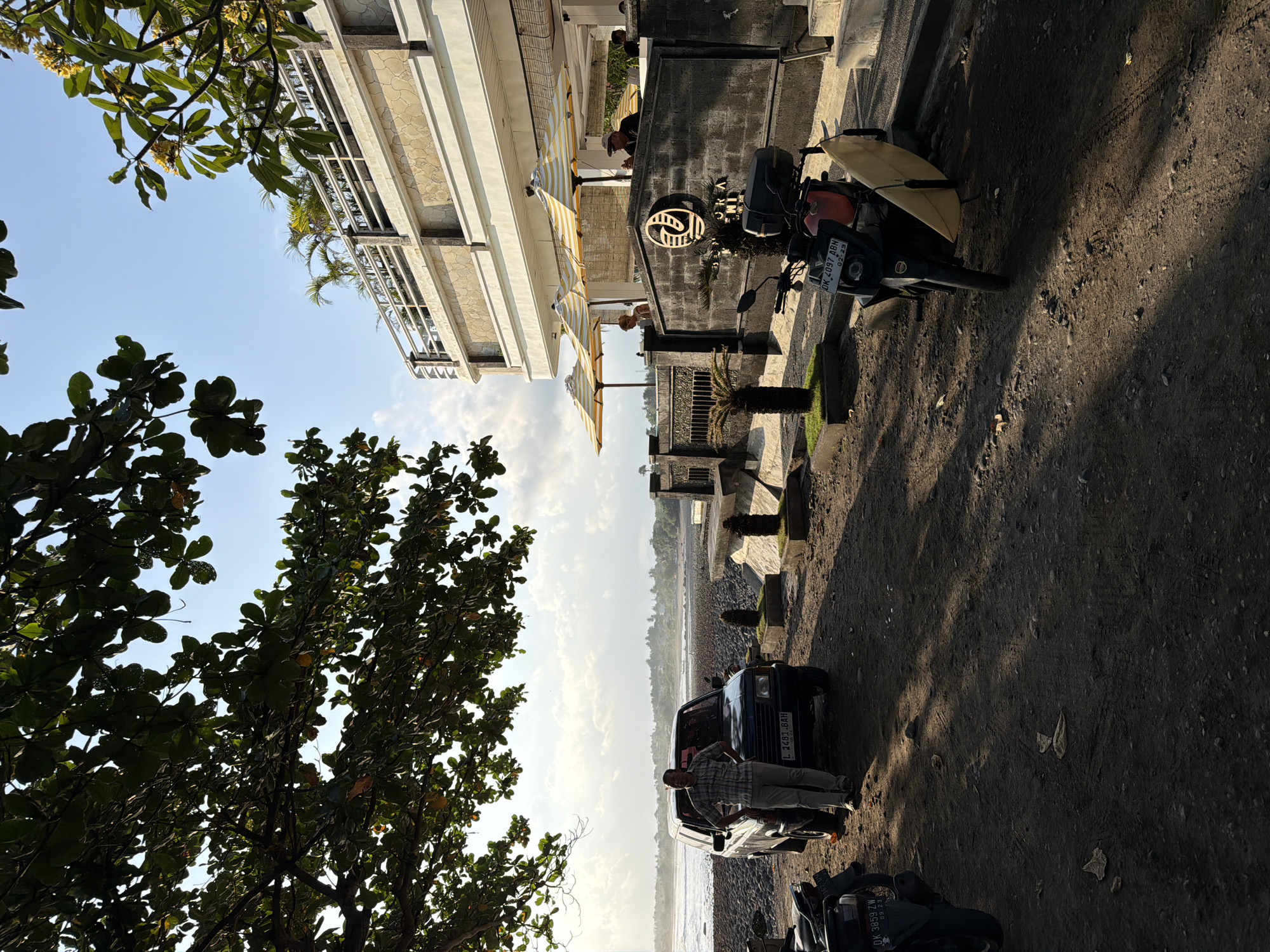 This is a medium shot of a beachfront scene in Pekutatan, Indonesia. In the foreground, a man stands next to a dark-colored SUV parked on a dirt and gravel lot. The man is wearing a plaid shirt and light-colored pants. A motorcycle with a surfboard attached is parked to the right of the SUV. In the background, a modern building with multiple levels and a balcony overlooks the ocean. Several umbrellas are set up on the balcony. The ocean is visible with a cloudy sky above. The ground is covered in dark soil and pebbles. Trees with green leaves are present in the upper left of the frame, casting shadows on the ground. The sun is low in the sky, creating long shadows.
