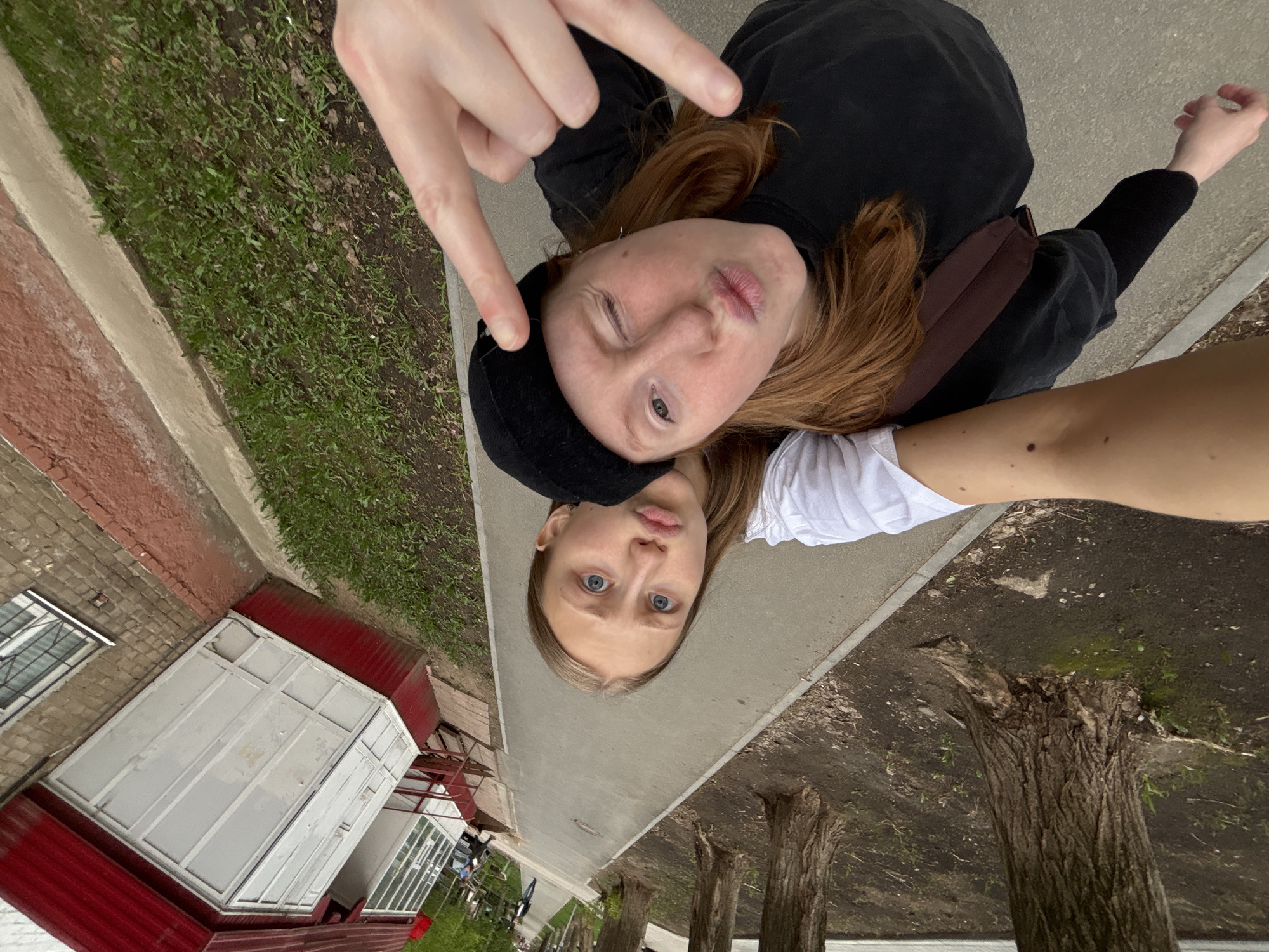 Two young women lie perpendicular to each other on a gray sidewalk. The woman at the top, with reddish-brown hair, wears a black hooded sweatshirt and a black hat, and makes a hand gesture with her left hand. The woman at the bottom wears a white shirt and has blond hair and blue eyes. A building with a red roof and white exterior and windows sits to the left of the sidewalk, and tree stumps and foliage line the right side. The location is Perm, Russia.