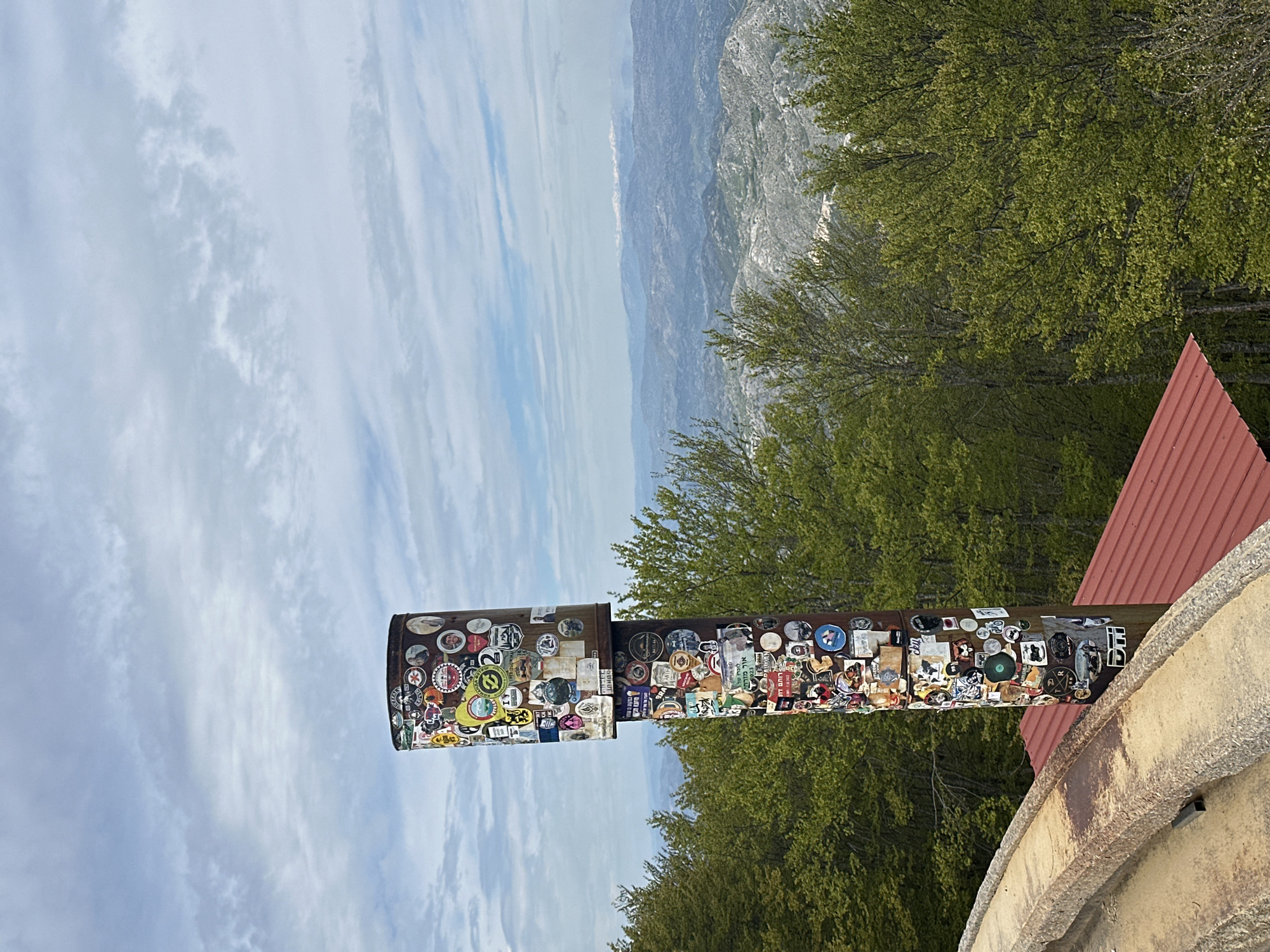 A low-angle, landscape-oriented shot depicts a sticker-covered metal pipe structure, possibly a viewing apparatus, situated on a weathered concrete ledge with a red, corrugated metal roof visible nearby. The view beyond includes a backdrop of lush green trees, and in the distance, a hazy mountainous terrain under a sky filled with scattered clouds. The location is implied to be in or near Mali Bostur, Montenegro. There are no people in the image.