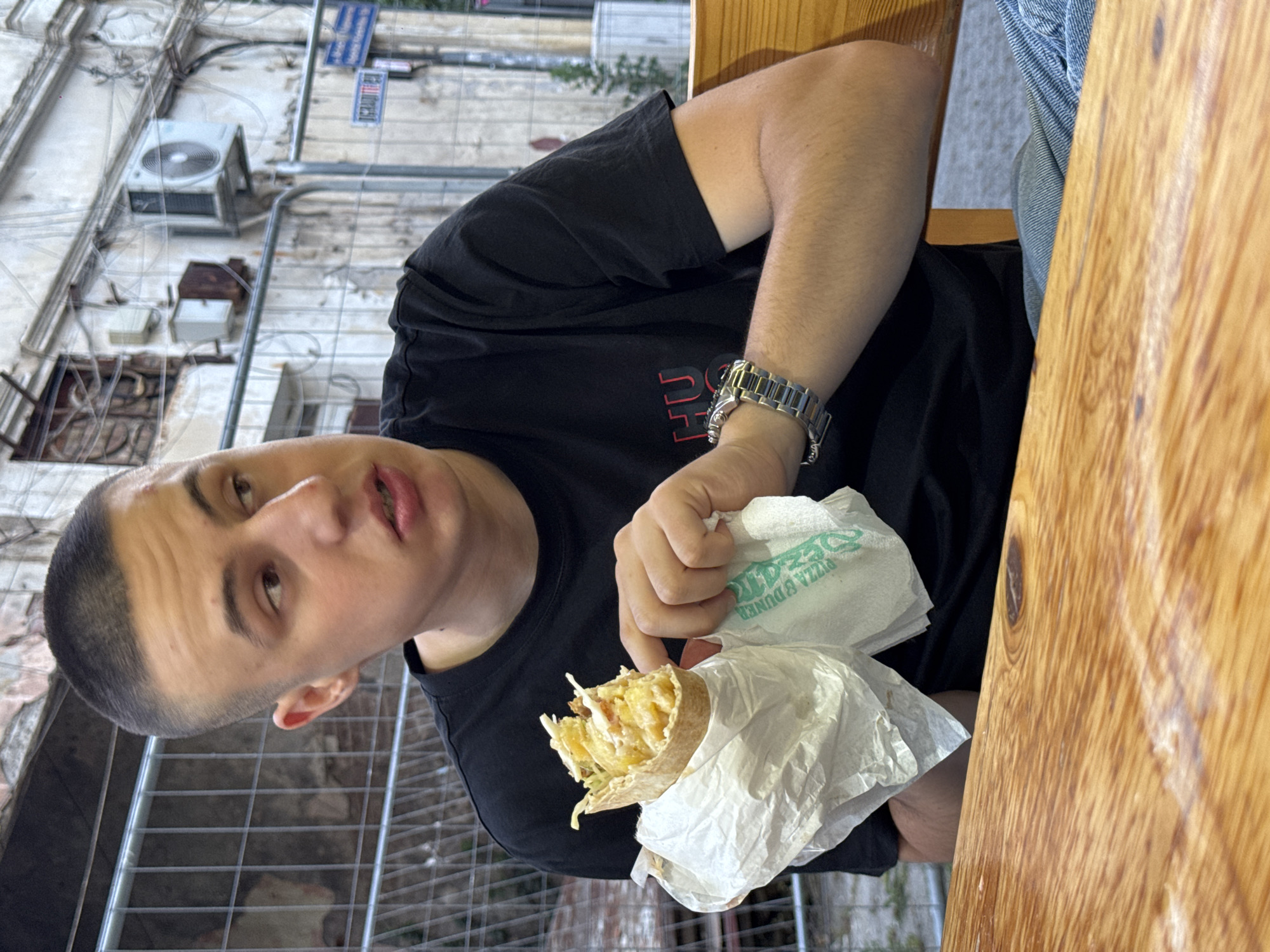 A young Caucasian male with a buzz cut, wearing a black t-shirt and jeans, is seated at a wooden table, holding a partially eaten wrap. He is looking off to the right of the frame with his mouth slightly open. A silver watch is visible on his left wrist. In the background, a building with visible air conditioning units and scaffolding is present. The scene appears to be an outdoor seating area, possibly at a food establishment, near a street in Burgas, Bulgaria.