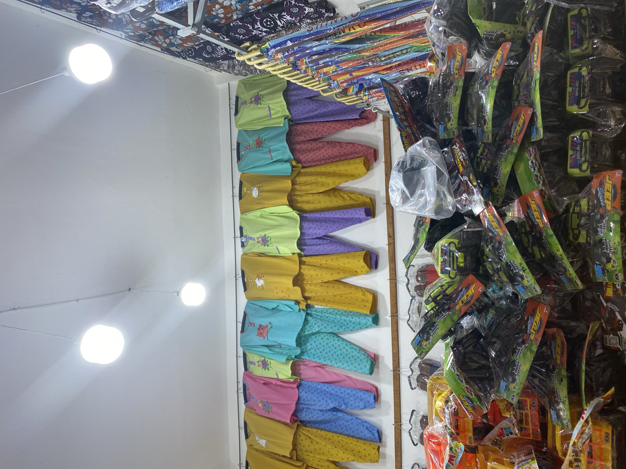 The image shows the interior of a shop in Lamteumen Barat, Indonesia. The shop is stocked with various items, including children's pajamas, toys, and umbrellas. In the foreground, there is a large display of toy vehicles, packaged in plastic. Behind this, a rack of colorful umbrellas is visible. The mid-ground features a display of children's pajamas, hung on hangers. The pajamas are in a variety of bright colors and patterns, some with cartoon characters. The background shows the ceiling with two bright, modern light fixtures. The overall impression is of a busy retail environment selling goods for children.