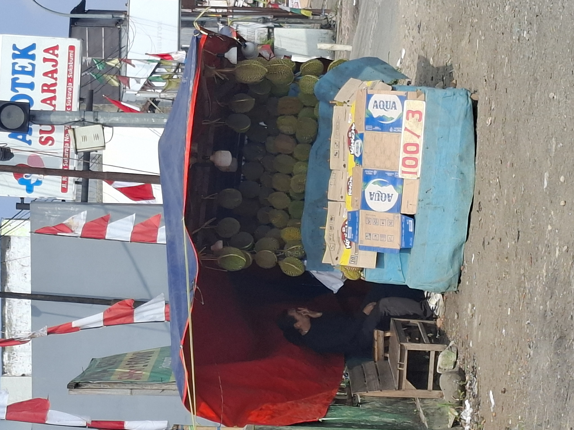 A woman sits on a wooden stool under a makeshift stall in Sukaraja, Indonesia. The stall is covered by a blue tarp and a red fabric, and it displays a large quantity of durian fruit hanging from the top. Boxes of Aqua bottled water and a sign that reads "100/3" are visible in front of the stall. In the background, Indonesian flags are displayed, and a sign for "Apotek Sukaraja" is visible on a building. The ground is dirt and debris, suggesting an outdoor market setting.