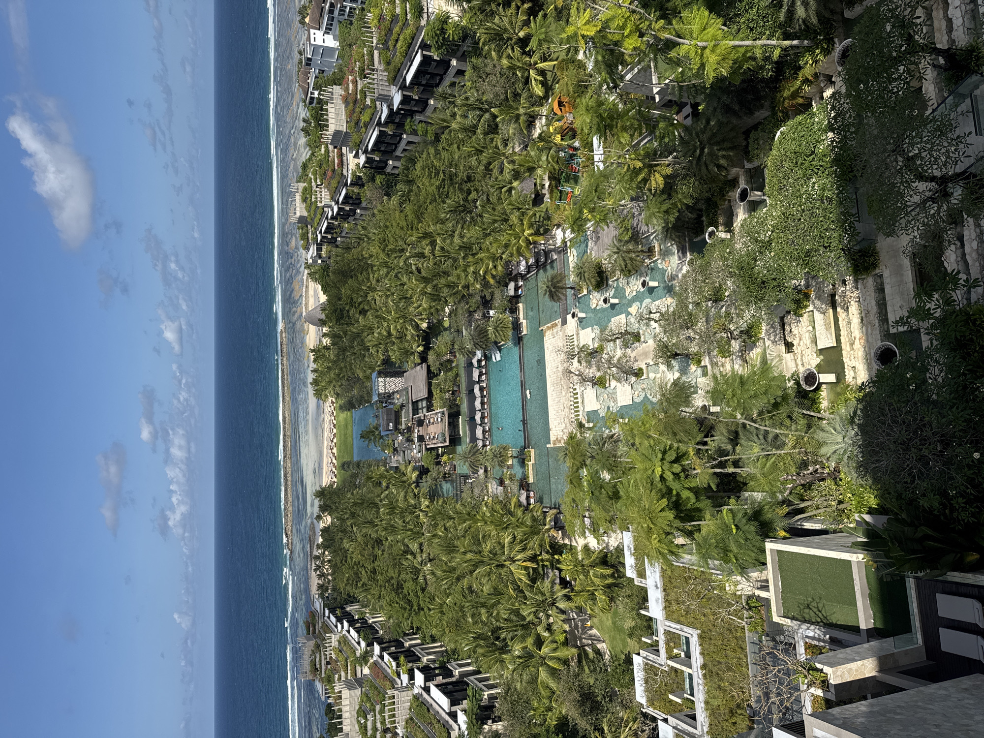This is an aerial view of a luxury resort along the coastline in Kuta Selatan, Indonesia. The scene captures the beach, the ocean, palm trees, swimming pools, buildings, and some vegetation. On the left side, there's a blue ocean with visible waves and a sandy beach. Adjoining the beach, the resort features multiple swimming pools, connected by walkways. Adjacent to the pools are sun loungers. There are many buildings with rooftop gardens. The predominant color in the image is green, due to the trees and vegetation covering the property. There is no visible activity, and the scene is serene.