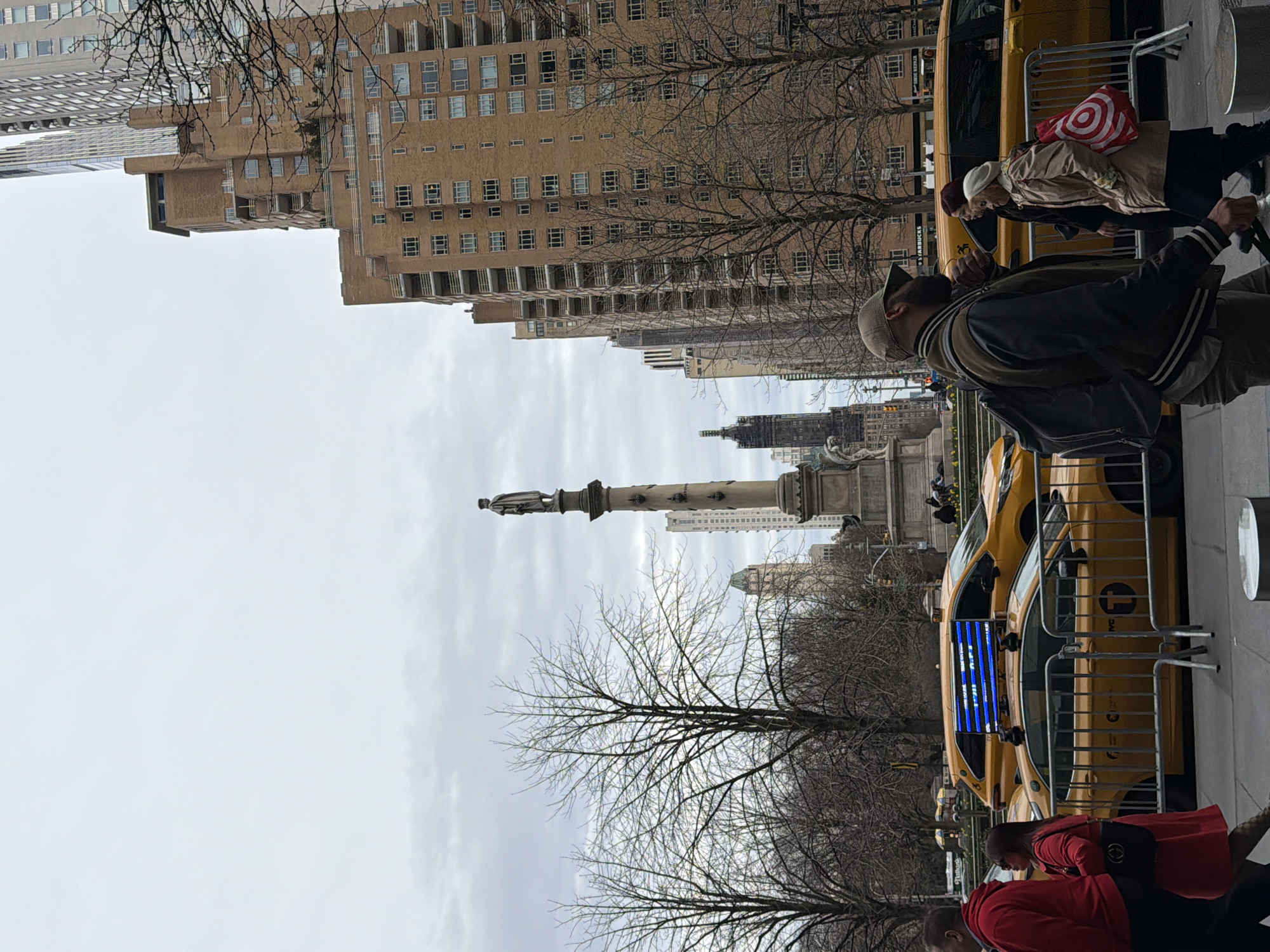The image displays an urban street scene at Columbus Circle, Manhattan, New York City, under an overcast sky. The Christopher Columbus Monument, featuring a statue atop a tall, stone column, is centrally located in the midground. To its left, several bare deciduous trees are visible, indicative of a park setting (Central Park). To the right, multi-story buildings, primarily constructed of light brown brick with numerous windows and balconies, line the street. Taller, modern skyscrapers are visible in the background, including structures consistent with the Deutsche Bank Center and other residential towers.
On the street level, several yellow New York City taxi cabs are present, some equipped with illuminated digital advertisements on their roofs displaying blue text. A metal barricade separates the sidewalk from the street traffic. Multiple pedestrians are walking on the sidewalk in the foreground and midground. In the immediate foreground, a male subject wearing a dark jacket, a baseball cap, and a backpack walks towards the left. Beside him, an elderly female subject in a beige coat and white beanie carries a red and white Target shopping bag while walking towards the right. A female subject with red hair and a dark jacket is partially visible behind them. On the far left, a male subject in a red hoodie and a female subject in a red coat carrying a black handbag are walking towards the right. The scene depicts a typical busy metropolitan environment.