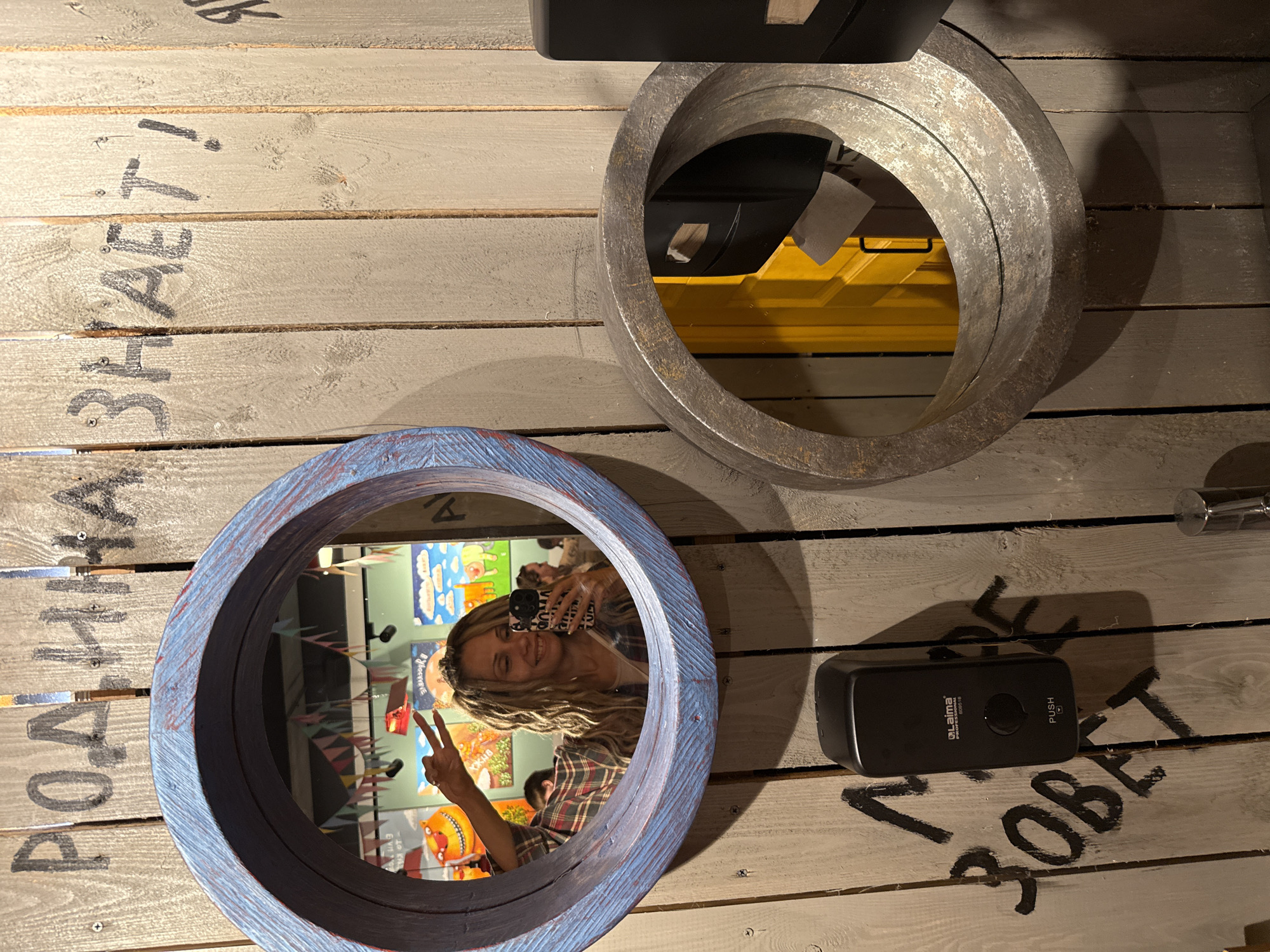 This media file captures an indoor scene in Заволжье, Russia, characterized by a wall or floor made of light-colored wooden planks. A prominent feature is a young woman, seen reflected in a circular mirror, taking a selfie. She is smiling cheerfully and holding up two fingers in a peace sign. She has long, wavy blonde hair and is wearing a plaid shirt, holding a smartphone with a patterned case.
Two circular frames are visible against the wooden background. The lower one, which reflects the woman, has a rustic, distressed blue and red painted finish. The upper frame is larger, appearing to be made of aged metal, and reflects a bright yellow surface along with some dark objects.
Black Cyrillic text is painted directly onto the wooden planks. On the left side, rotated, the phrase "РОДИНА ЗНАЕТ!" (The Motherland knows!) is discernible. On the right, below a black wall-mounted dispenser, the word "ЗОВЕТ" (calls) is painted, suggesting it might be part of the patriotic slogan "РОДИНА ЗОВЕТ" (The Motherland calls!). The black dispenser itself bears the text "Laima PROFESSIONAL SDG-101" and "PUSH".
The mirror's reflection also reveals colorful, possibly artistic, elements in the background, including text that appears to say "DISCOVER!" and "OCKER". The lighting suggests an indoor setting, and shadows from the dispenser and upper frame are cast on the wooden surface. The overall impression is one of a lively, possibly themed, interior space.