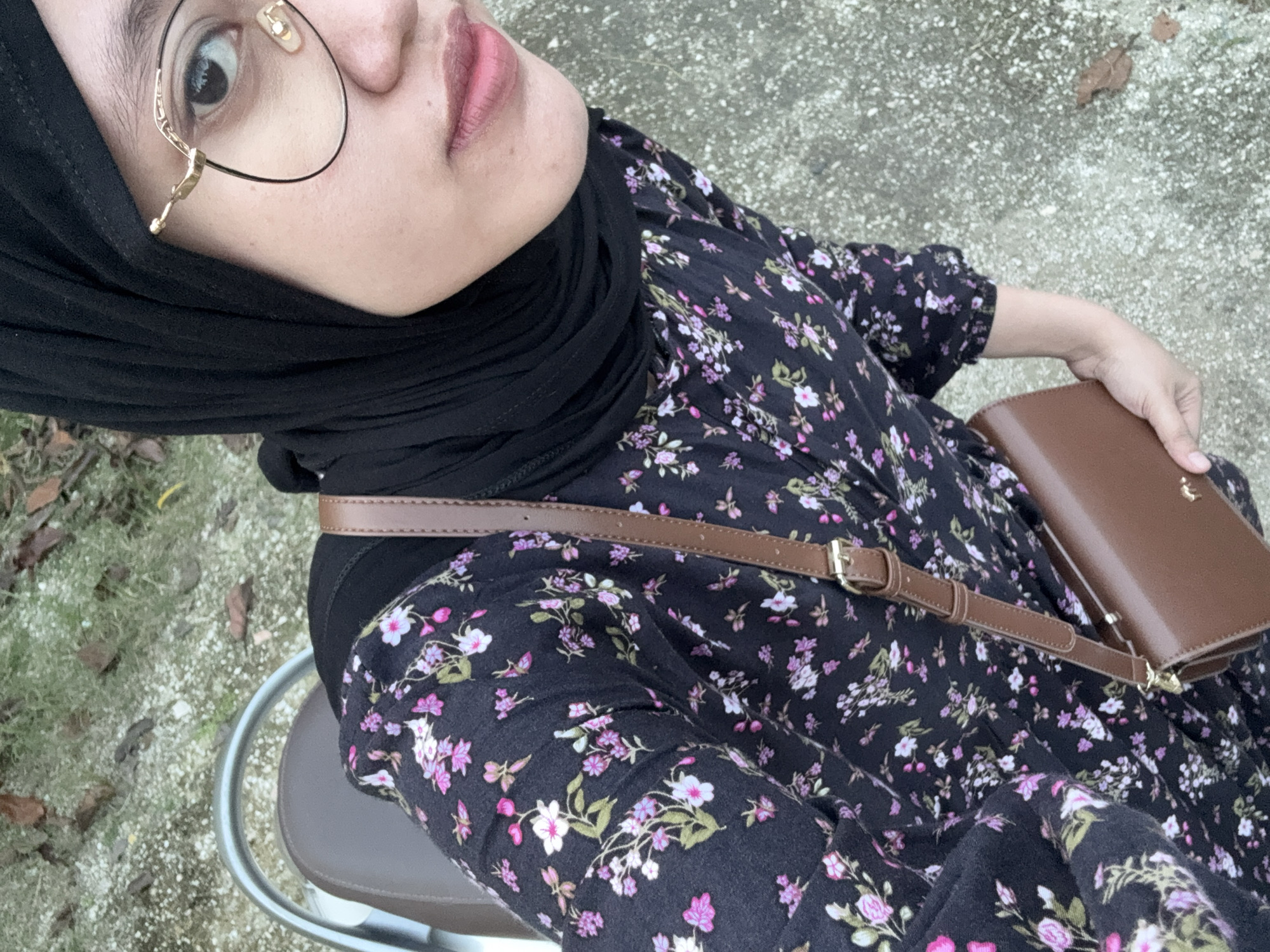 A woman is seated outdoors in Gadingrejo, Indonesia. She wears a black hijab, round gold glasses, and a long-sleeved dress with a dark floral pattern. She has a brown leather purse with a matching shoulder strap. The ground is light-colored gravel with patches of greenery. The woman is sitting on a stool with a silver frame and a tan seat.