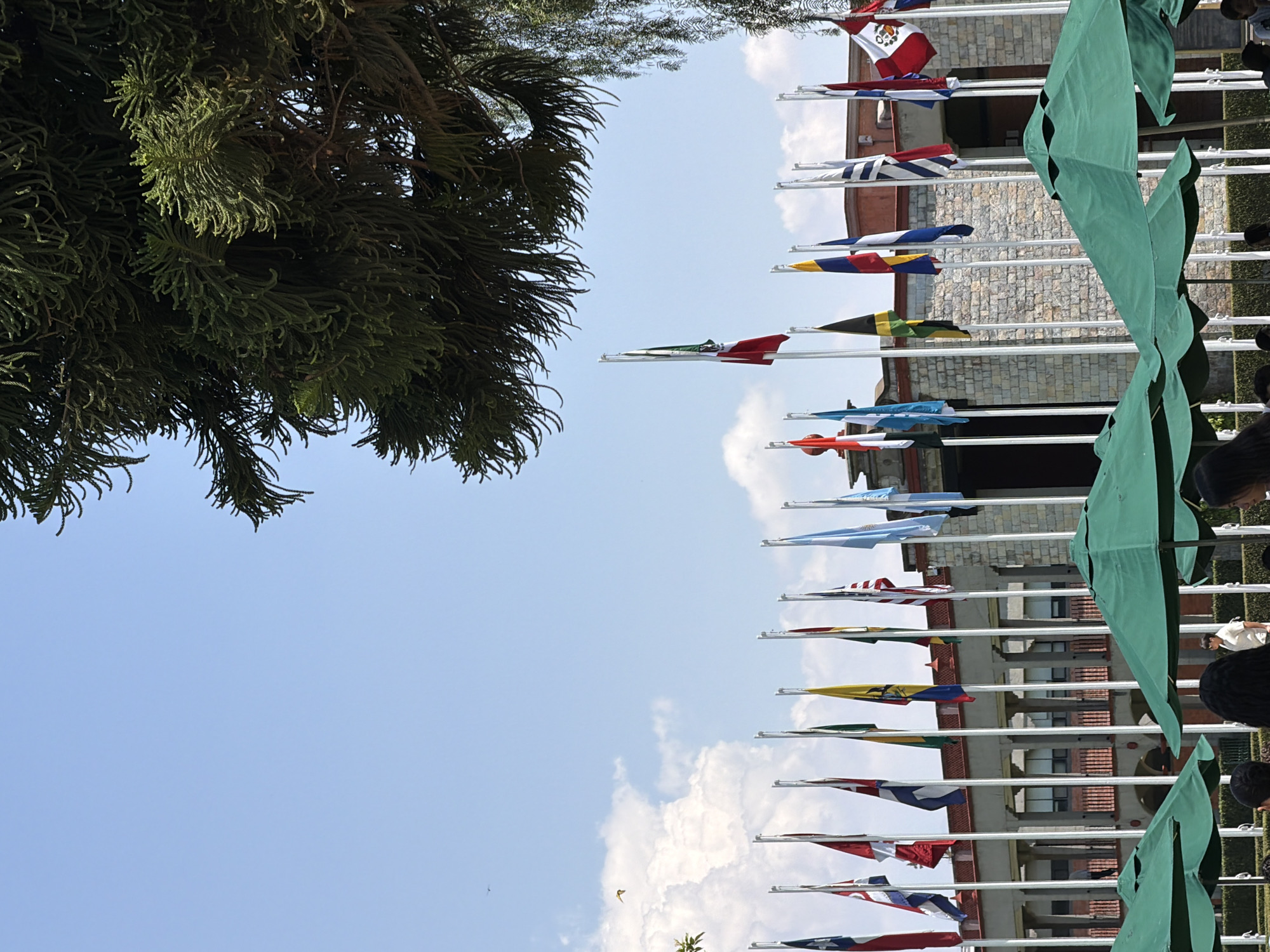 Here is a detailed description of the image:
The image presents a collection of flags representing different nations, likely placed in front of a building, possibly a government facility or an international institution. The flags are attached to individual poles, positioned closely together in a row. To the right, there are green canopies and people. To the left, there's a tree with dense foliage. The sky is visible in the background, with patches of clouds. The flags, canopies, and people are in focus, while the tree is slightly out of focus. The building in the background is a multistory structure with a stone facade and multiple windows. The exact location of the image cannot be determined because the city and country are unknown.