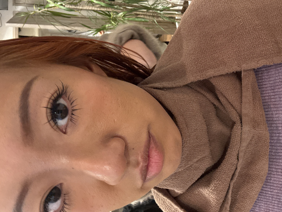 A close-up, head-and-shoulders portrait of a young woman with a medium complexion and reddish-brown hair. She gazes directly forward, featuring defined eyebrows, dark eyes with pronounced, long eyelashes, and subtly blushed cheeks. Her nose and full lips, slightly parted, are visible. A brown terry cloth towel is wrapped snugly around her neck, covering a light purple, ribbed garment. The background is softly out of focus, revealing an indoor environment with several tall green potted plants. A blurred figure, showing a light-colored, possibly fur-trimmed garment on a shoulder and arm, is partially visible behind the subject's right side. The scene is set in an establishment in Toshima, Japan, consistent with a beauty salon or similar personal care venue.