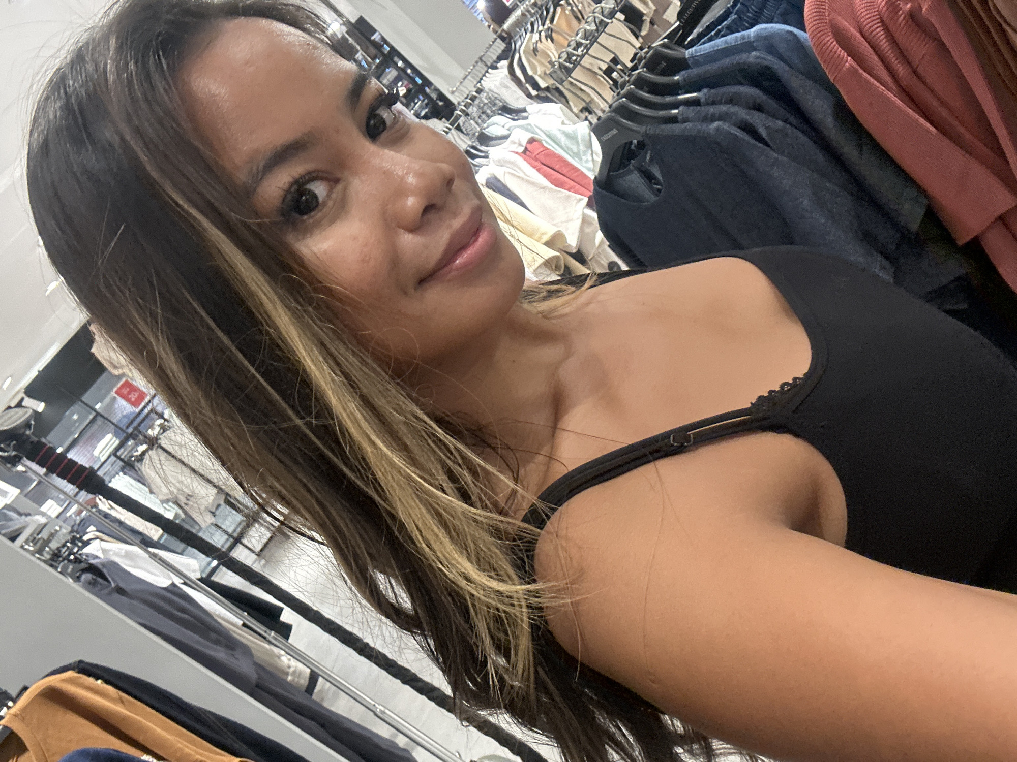 The image is a selfie taken in a clothing store, likely in Denpasar, Indonesia. A woman with long, wavy brown hair with blonde highlights is in the foreground, smiling at the camera. She is wearing a black tank top. Behind her, racks of clothing are visible, including shirts and sweaters in various colors and styles. The lighting suggests it is indoors during the day.