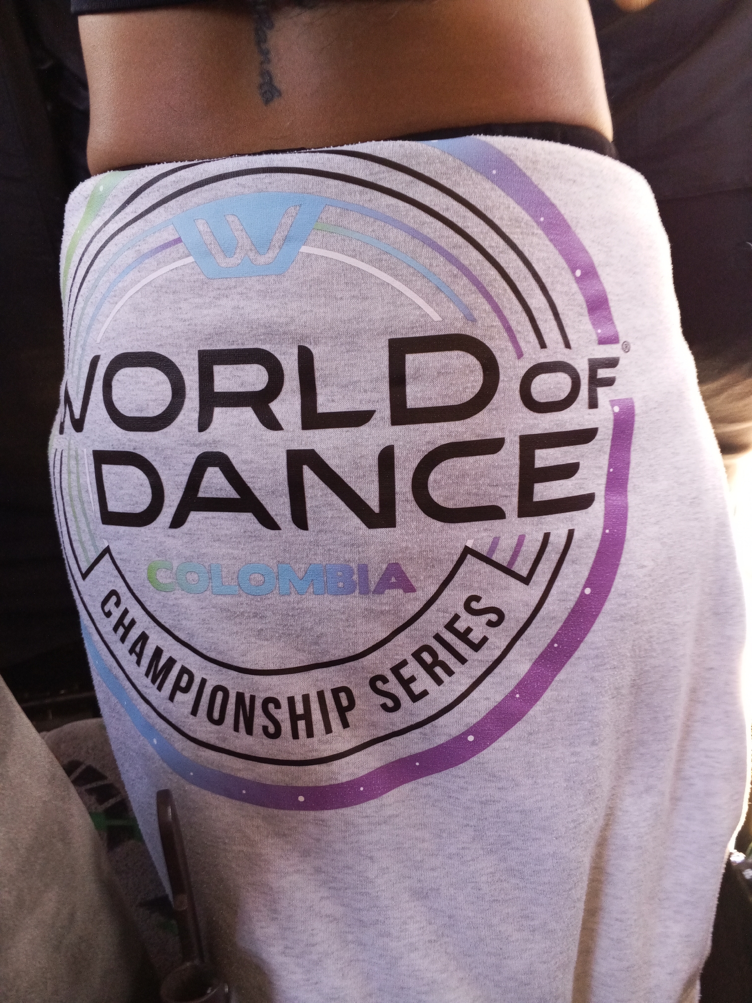 A close-up shot features the lower back and upper hip area of a person, revealing a light grey garment adorned with a prominent graphic design. The background is dark and softly blurred, suggesting an indoor setting or a shaded outdoor area.
The primary subject is the light grey fabric, possibly sweatpants or shorts, which features a large, colorful print. The print consists of a circular logo with the bold black text "WORLD OF DANCE" centrally displayed. Below this, the word "COLOMBIA" is spelled out in smaller, gradient letters, transitioning from green to light blue and purple. Curved along the bottom arc of the circle, the words "CHAMPIONSHIP SERIES" are written in black capital letters. At the top of the design, a stylized light blue "W" logo is encased in a hexagonal shape, accompanied by concentric arcs in light blue, light purple, and dark purple, some dotted with small white specks. A small registered trademark symbol is visible near the word "OF".
Just above the garment, on the exposed skin of the person's lower back, a dark, vertical tattoo with illegible script is visible. The lighting appears to be natural and bright, highlighting the texture of the fabric and the details of the print, suggesting it is daytime. A dark strap or handle of an unseen object is partially visible in the bottom-center of the frame. No specific activity is depicted other than the person wearing the branded apparel.