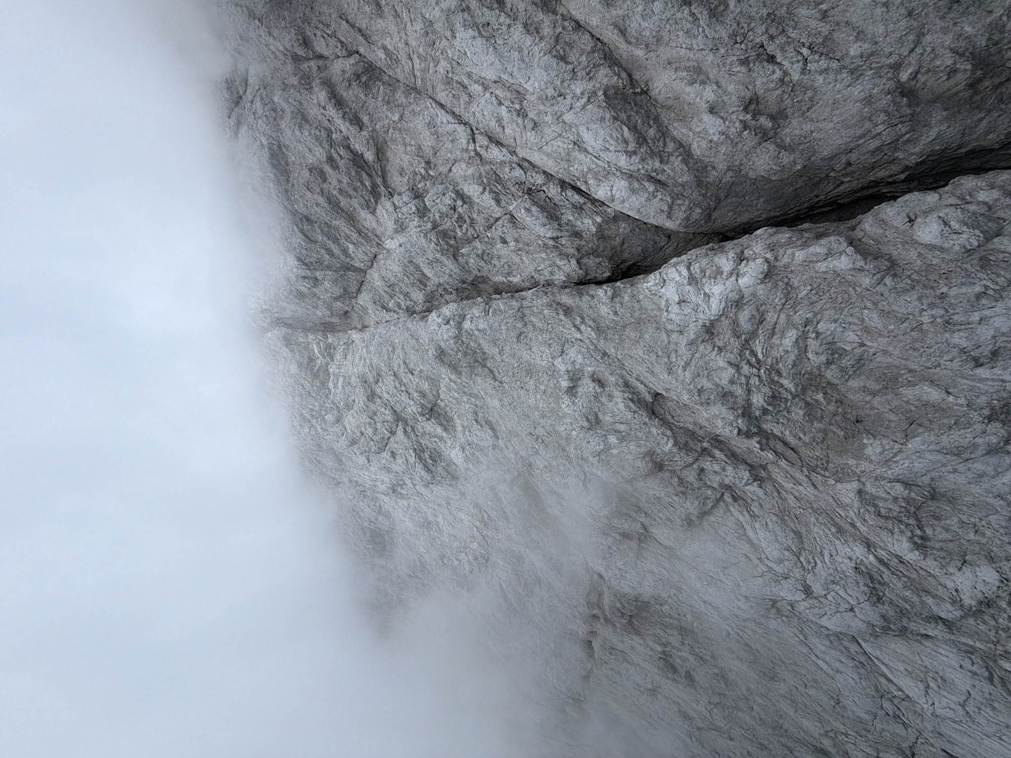 This media file is a close-up, top-down view of a steep, gray rock face partially obscured by a thick, white fog or cloud. The rock surface is textured and rugged, with visible fissures and cracks, suggesting a natural, mountainous environment. The fog creates a soft, diffused light and obscures the lower and left portions of the rock face, giving a sense of depth and mystery. The scale is difficult to determine without any reference points, but the texture of the rock implies a large, geological formation.
There are no people or objects visible in the frame. The scene depicts a natural, atmospheric landscape. The time of day is indeterminate due to the overcast conditions, but the diffuse light suggests it is daytime. The weather is clearly foggy or cloudy, creating a dramatic and somewhat somber mood. The location context provided, Grainau, Germany, indicates a mountainous region, likely the Alps, known for its dramatic rock formations and frequent fog. There is no visible text in the image.