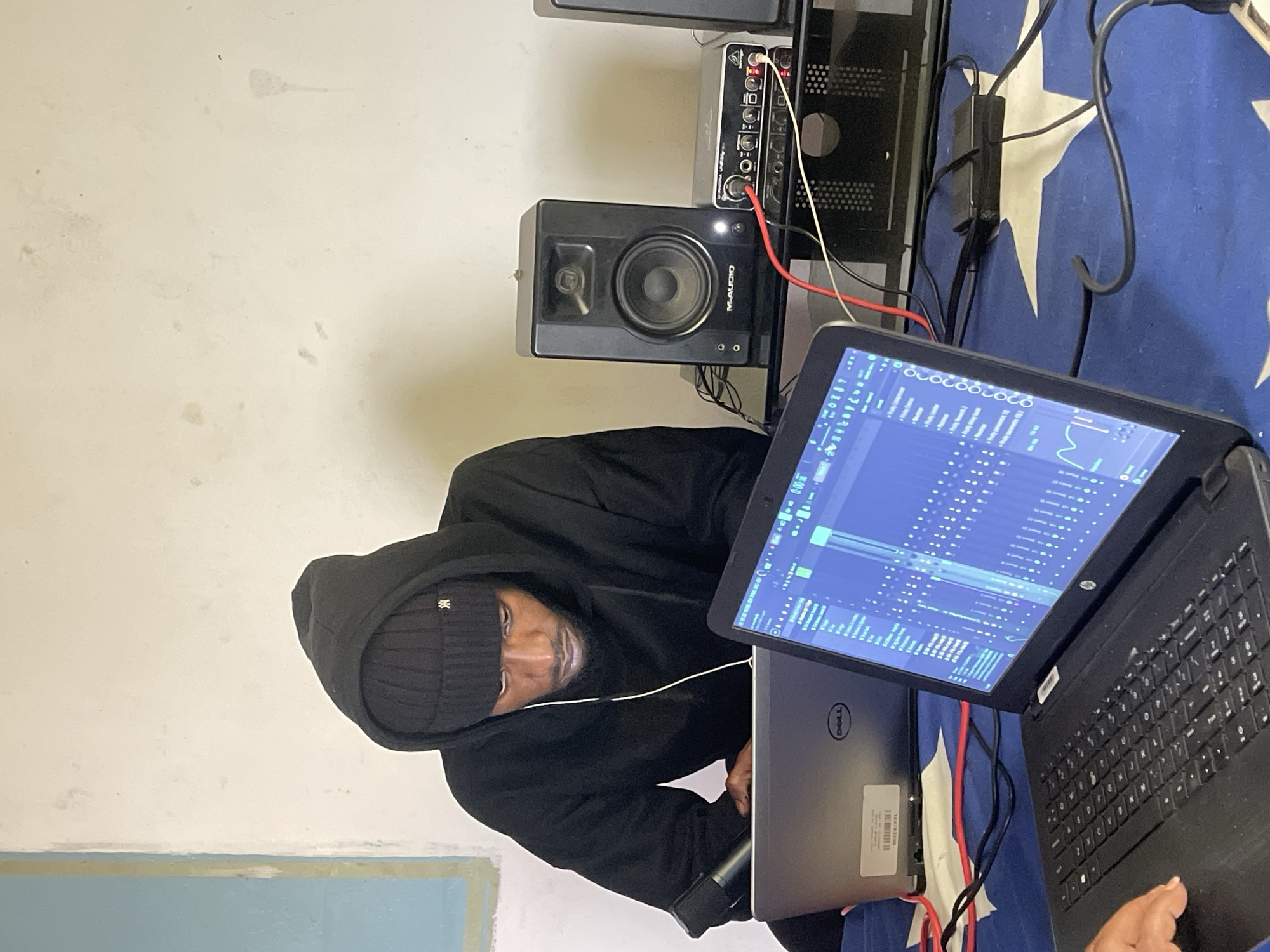 A Black male wearing a black hoodie and a black beanie is seated at a desk in a room. He is looking down at two laptops that are positioned in front of him. The laptop on the right displays a digital audio workstation (DAW) interface with a complex arrangement of tracks, waveforms, and control elements, indicating music production or audio editing. A black studio monitor, labeled "M-AUDIO," is visible to the right of the laptops, along with an audio interface labeled "BEHRINGER UMC204HD" and other electronic equipment. The setting appears to be a home studio or a creative space. The wall behind the subject is a light, textured color, with some blue and yellow paint visible on the left side, potentially indicating a window frame or wall decoration. The desk surface is covered with a blue cloth featuring a white star pattern. The scene suggests a focused and engaged individual involved in music production. The presence of specific brands like Dell and HP on the laptops, along with M-Audio and Behringer audio equipment, suggests a functional setup for audio work. The lighting in the room appears to be artificial, casting some shadows. The overall environment suggests a working studio space, possibly located in Jos, Nigeria, given the prompt's constraint, though there are no definitive visual cues within the image to confirm this specific location.