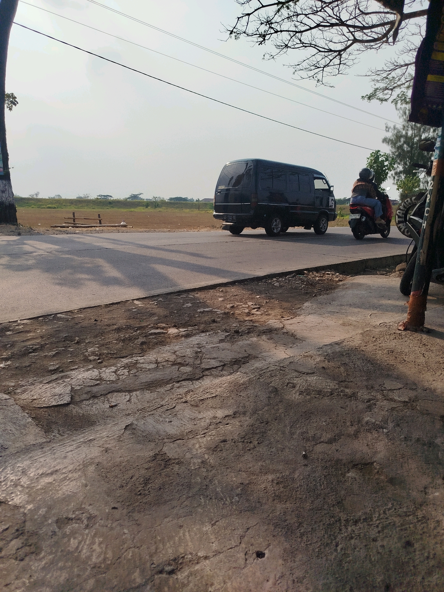 In the city of Ngreco, Indonesia, a black van is traveling down a paved road. To the right of the van, a person is riding a red motorcycle. The road is bordered by a dirt path and sparse vegetation. The sky is clear and bright, suggesting it is daytime. The scene is captured from a slightly elevated perspective, looking down on the road and surrounding area.