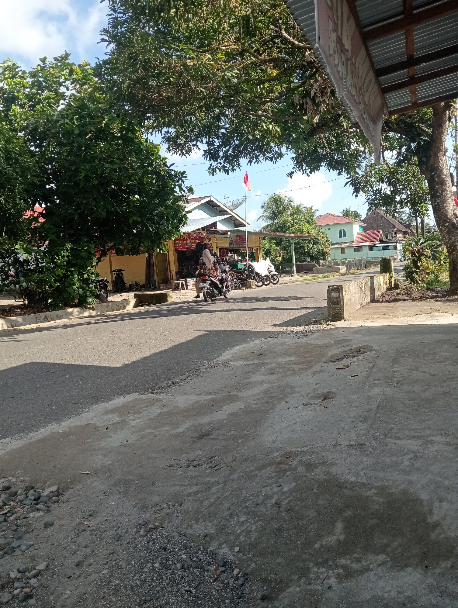 The image depicts a street scene in Campago, Indonesia. On the right side of the frame, a concrete structure, possibly a low wall or bridge abutment, is visible. Beyond it, lush green trees and vegetation line the area. A large tree trunk with rough bark is prominent on the far right. The street itself is paved asphalt, with a lighter concrete or gravel shoulder on the right. On the left, a large, leafy tree casts a shadow across the road. A line of buildings with various roof types, including corrugated metal and red tiles, is situated along the left side of the street, set back from the road. One building features a yellow facade and a metal awning. Several parked motorcycles are visible in front of the buildings. In the mid-ground, a person wearing a headscarf and patterned clothing is riding a motorcycle, with another motorcycle behind it. Further in the distance, more buildings and palm trees are visible under a bright blue sky with scattered clouds. A red and white flag is flying on a pole in front of one of the buildings. The overall scene suggests a typical day in a semi-urban or suburban area of Indonesia.