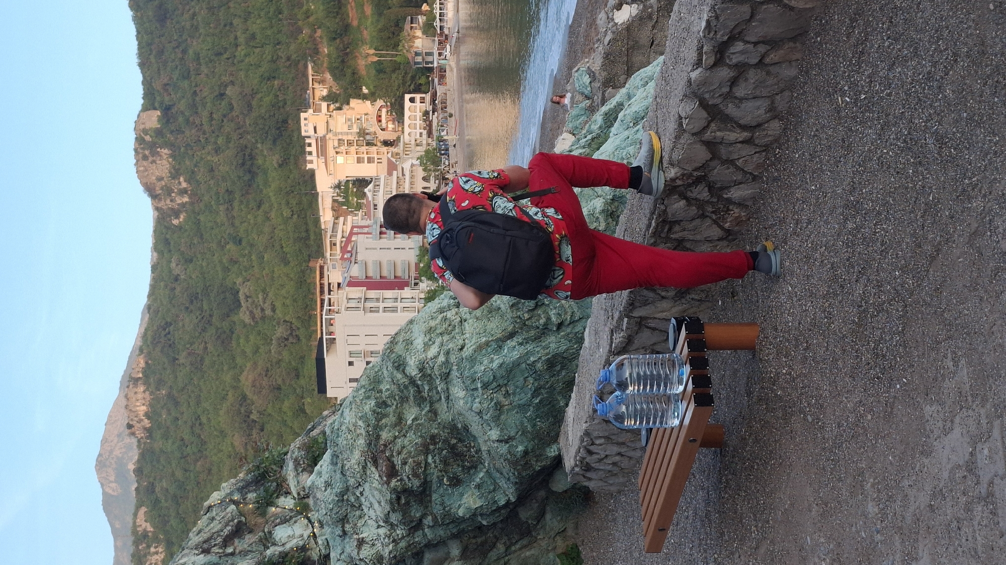The image captures a scene in Rafailovići, Montenegro, featuring a person sitting on a rocky ledge overlooking the sea. The individual is wearing a red outfit with a patterned shirt and a black backpack. They appear to be using a mobile phone. Two water bottles are placed on a wooden bench near the person. In the background, buildings and a beach stretch along the coast, with a green mountain visible on the left side of the frame. The landscape suggests a coastal Mediterranean setting.