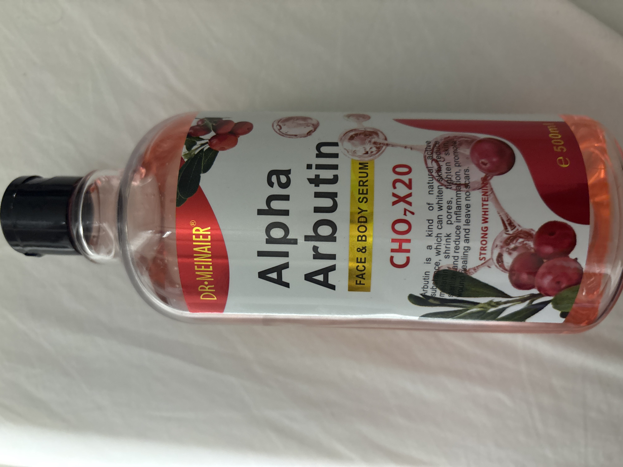 The image shows a close-up, slightly angled, full-body shot of a clear plastic bottle with a black cap, filled with a pinkish liquid. The bottle has a label with the brand name "DR. MEINAIER" in gold lettering on a red background. Below this, the product name "Alpha Arbutin" is printed in large, black, serif font. Further down, in a yellow rectangle, it says "FACE & BODY SERUM". Below that, in red lettering, are the characters "CHO,X20". A descriptive text in smaller black font states "Arbutin is a kind of natural active substance, which can whiten skin, reduce melanin, shrink pores, tighten skin, sterilize and reduce inflammation, promote wound healing and leave no scars." Underneath this, in white lettering on a red background, it says "STRONG WHITENING". At the bottom of the label, in yellow lettering, it reads "e 500ml". The bottle is placed on a white, textured surface that appears to be fabric, possibly a bedspread or a blanket. The background is a blurred white, suggesting a domestic indoor setting. The lighting is soft and even, with no harsh shadows. There are no people or animals present in the image. The scene does not provide any specific geographical clues to indicate a location in Dubai, United Arab Emirates.