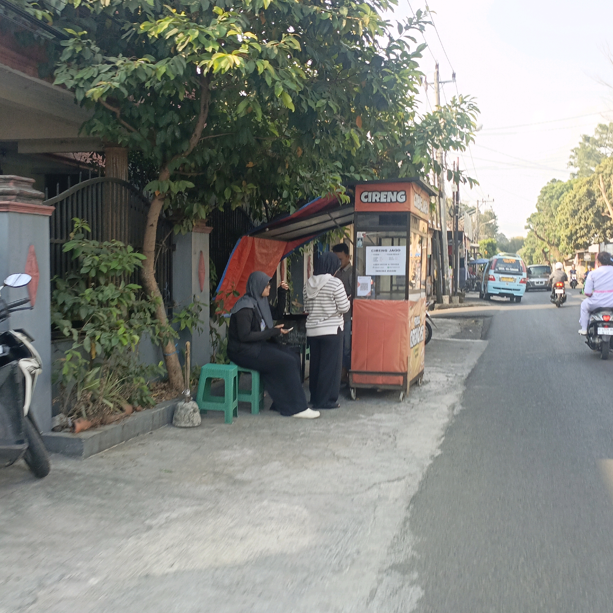A street scene in Salatiga, Indonesia, features a roadside food stall selling "Cireng Jago." Two women are at the stall; one, wearing a black hijab and long black dress, sits on a green plastic stool, holding a phone. The other, in a striped hoodie and dark pants, stands near the counter. A man is visible behind the counter, possibly an employee. The stall itself is orange and has signage. A motorcycle is parked to the left of the frame. The street is paved, with trees and buildings lining the side. Several vehicles, including a blue van and motorcycles, are on the road. The lighting suggests it is daytime, possibly late afternoon.