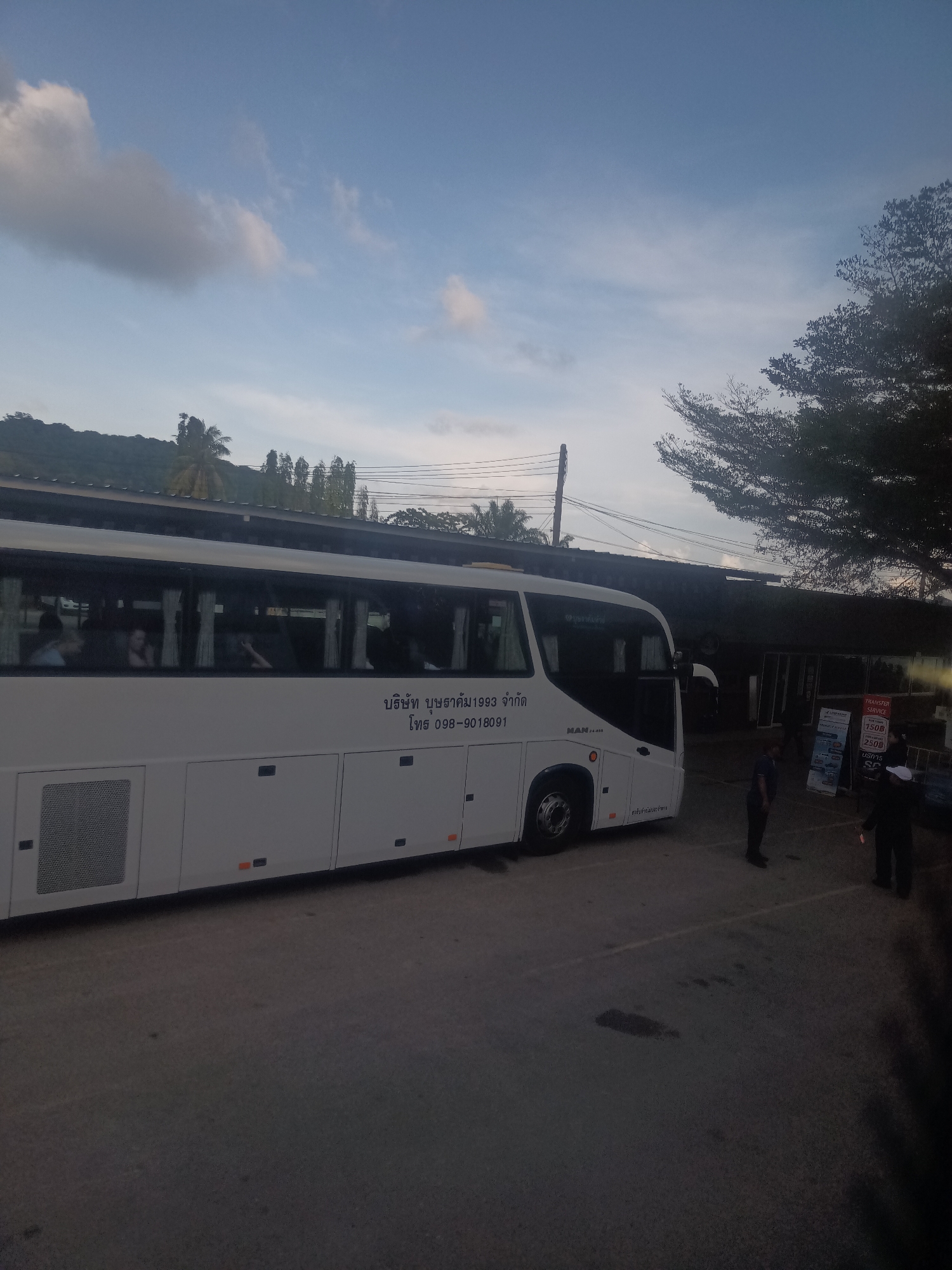 The image depicts a white MAN bus parked in an outdoor parking area in Tambon Hat Sai Ri, Thailand. The bus has Thai text and a phone number (098-9018091) displayed on its side. Several people are visible near a stand displaying "TRANSFER SERVICE" in English and Thai. The area appears to be a transportation hub or terminal. The sky is partly cloudy, and there are trees and buildings in the background.