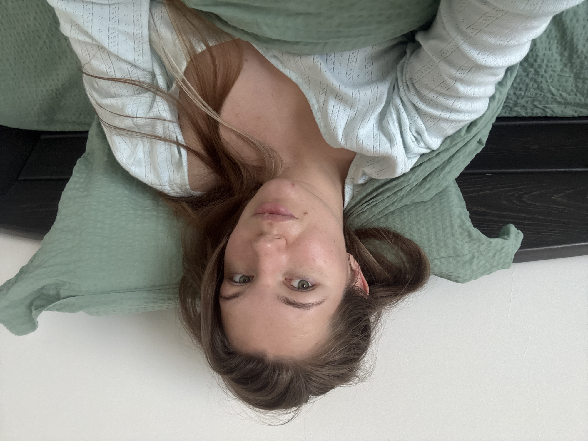 The scene is located in a room within the Vozdovac Urban Municipality, Serbia. A single female subject is positioned in a supine orientation, propped against bedding. She possesses medium-length brown hair, green eyes, and fair skin with visible freckles and a minor blemish on the chin. Her expression is neutral, and she gazes directly into the camera lens. She is attired in a light blue, long-sleeved garment featuring a textured, openwork pattern and a V-neckline. The subject is situated on a bed with dark green, textured pillows and a matching dark green blanket or duvet cover. A dark brown or black wooden headboard is partially visible on both sides of the subject's head. The background consists of a plain white wall. The lighting is soft and ambient. The perspective suggests a self-portrait.