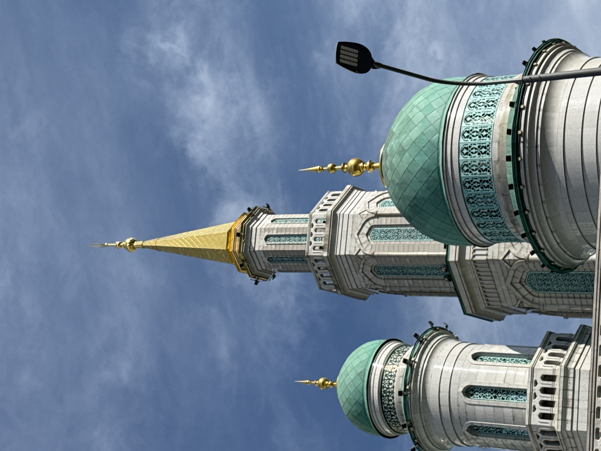 The image captures the intricate architecture of the Moscow Cathedral Mosque in Moscow, Russia, against a backdrop of a cloudy blue sky. The mosque features multiple minarets and domes, with the central minaret reaching highest, topped with a golden spire. The minarets are adorned with decorative turquoise accents and arched windows. In the foreground, a modern street lamp is visible, its dark silhouette contrasting with the lighter tones of the mosque and the sky. The scene is devoid of people, focusing entirely on the structure and its immediate surroundings. The overall impression is one of grandeur and detailed craftsmanship.