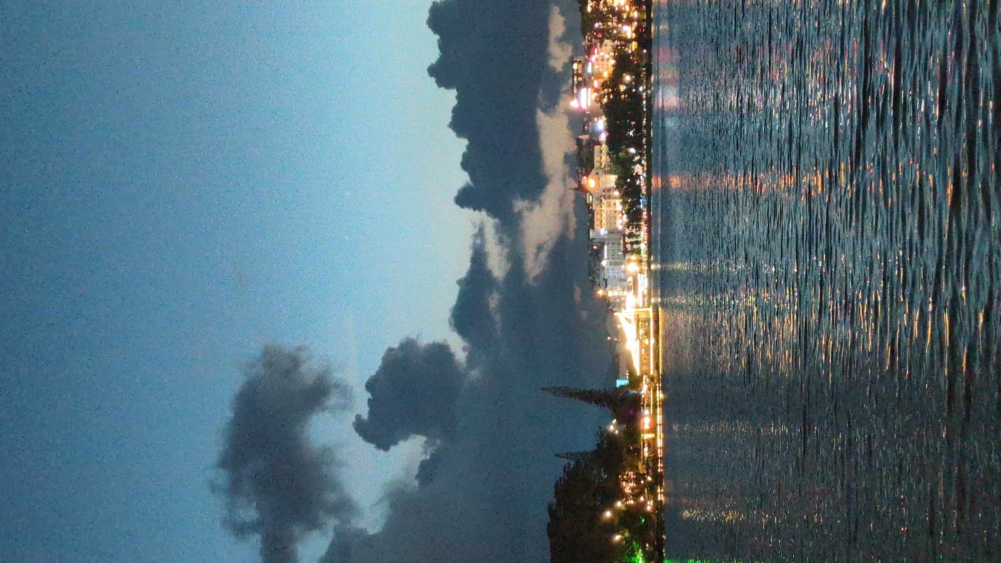 This is a nighttime photograph of Xuan Huong Lake in Da Lat, Vietnam. The sky is a deep blue with dark clouds scattered across it, creating a dramatic effect. The city skyline is visible in the background, illuminated by numerous lights from buildings and streetlamps. The lights are reflected in the calm waters of the lake, creating a shimmering effect. A few trees and structures are also visible along the shoreline, including what appears to be a long, illuminated building or boat on the left side of the frame, possibly a restaurant or tour boat. The overall atmosphere is serene and picturesque, with the vibrant city lights contrasting against the darkening sky and the tranquil water.