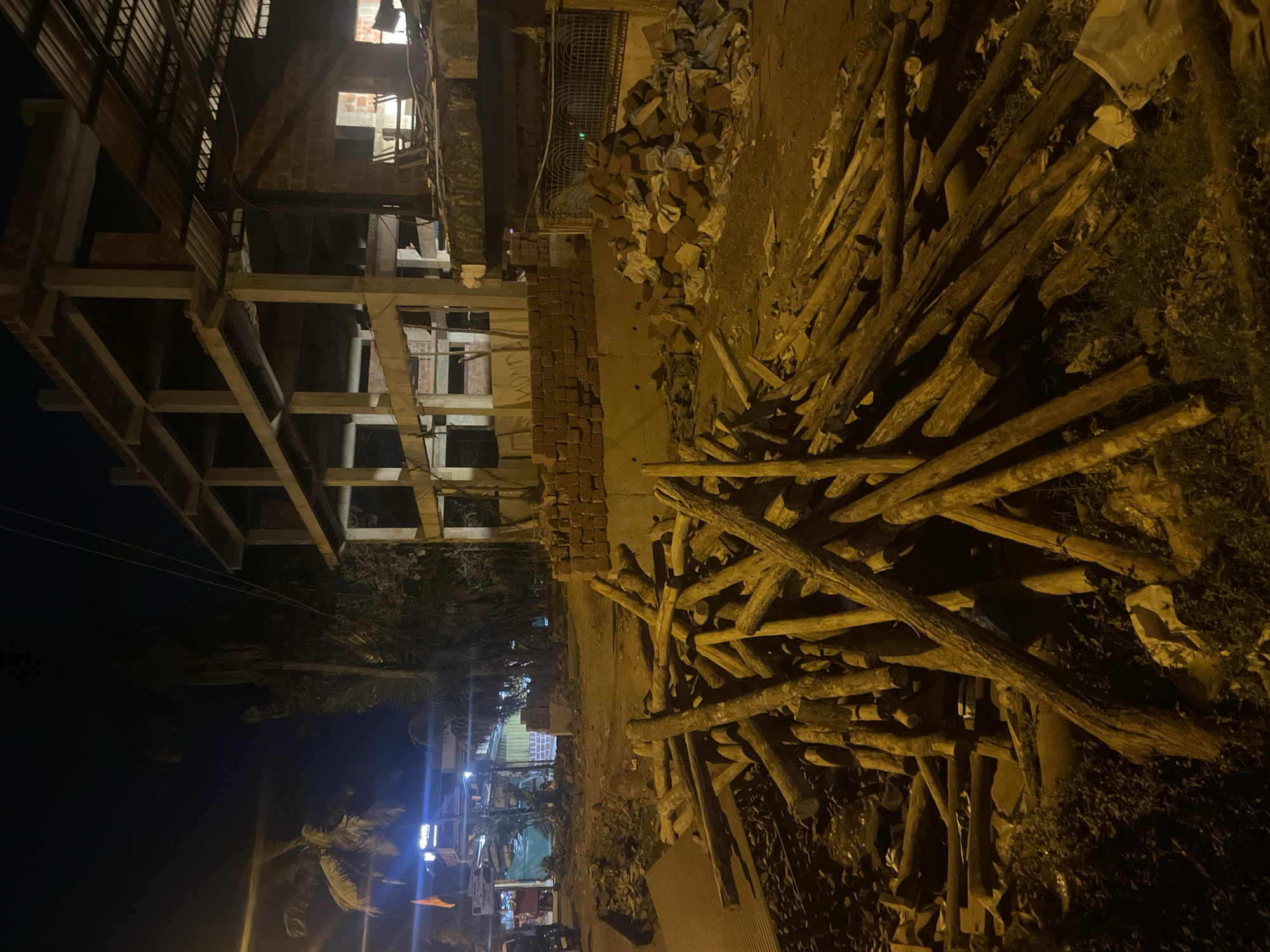 At night in Kudal, India, a partially constructed building stands next to a pile of logs and bricks. The building's concrete skeleton is visible, with a few brick walls. A pile of rough-cut logs occupies the foreground. Adjacent to the logs are stacks of red bricks. Streetlights illuminate a distant street scene with vehicles and shops.