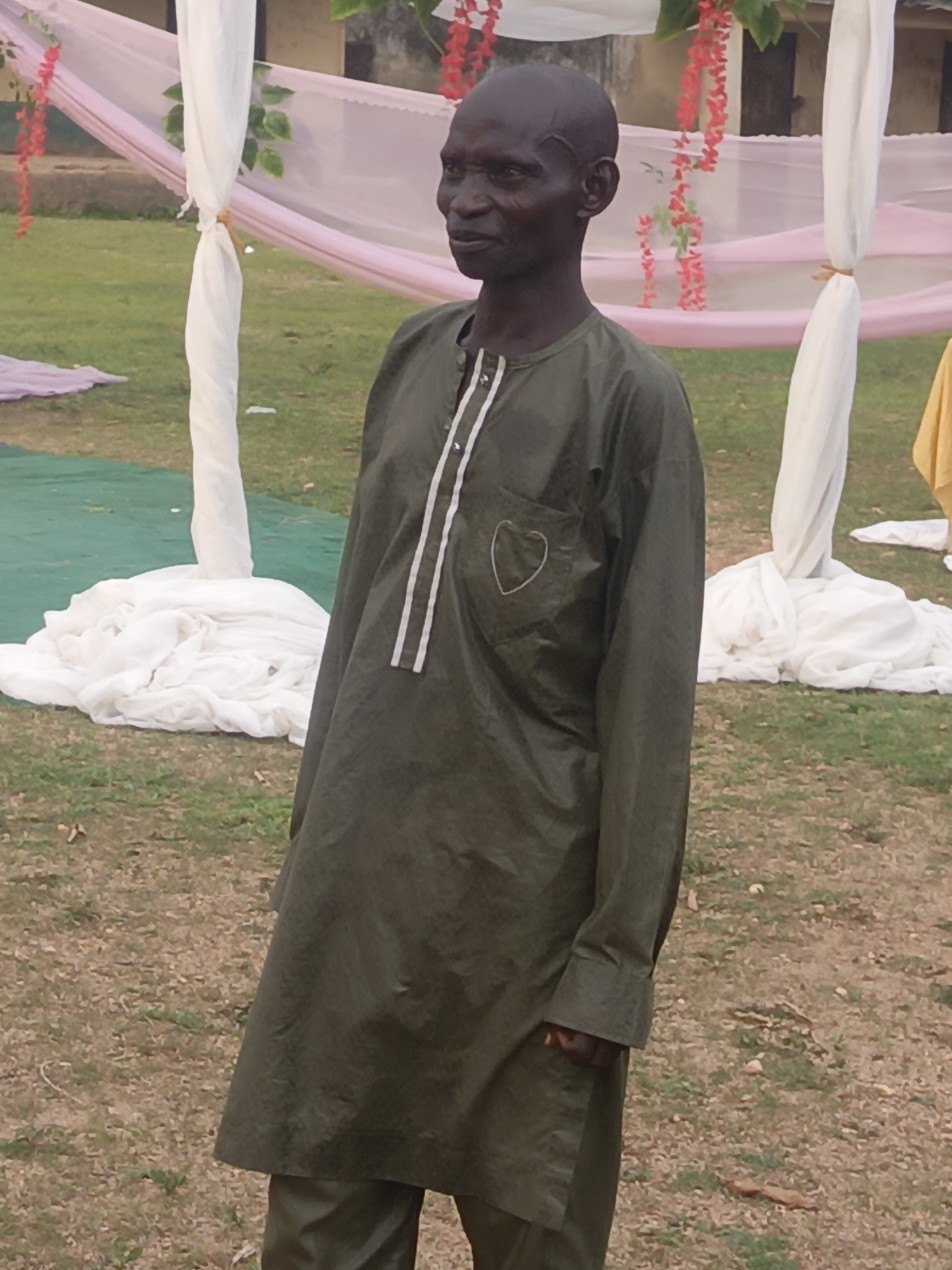 A man stands in an outdoor setting with decor that suggests a wedding or celebration. He is wearing a long-sleeved, olive-green tunic with white stripes down the front and a heart design on the chest pocket, along with matching pants. He is bald, with dark skin, and appears to be looking off to the side. The background consists of white and pink draped decorations supported by poles. The ground appears to be grassy with patches of bare earth. Based on the provided information, the location is in Unguwar Zaria, Nigeria.